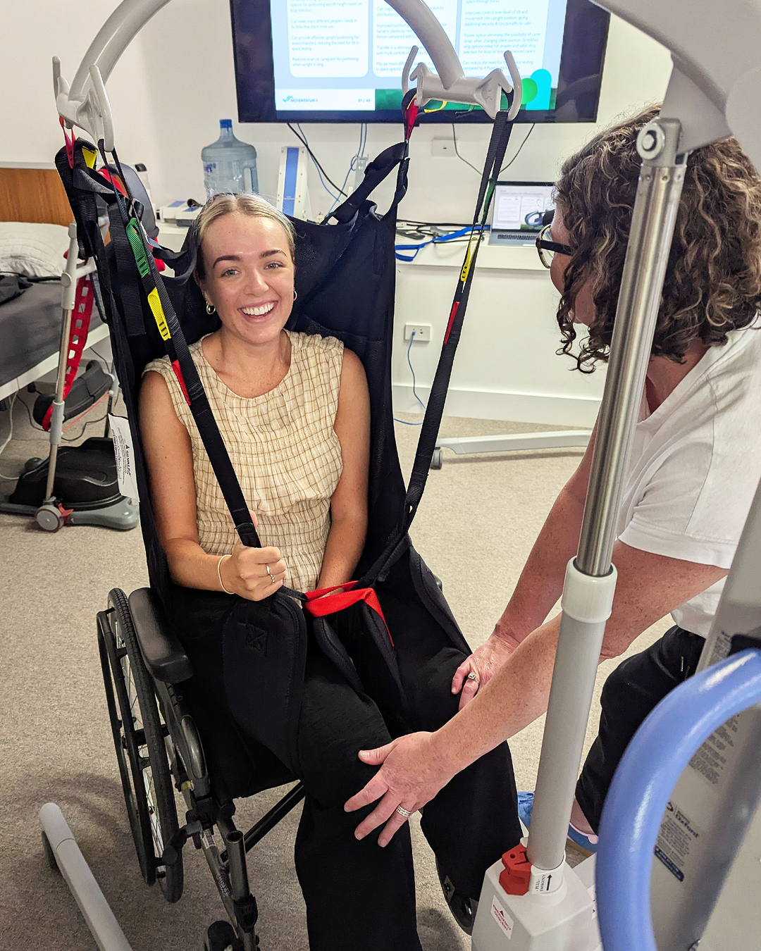 A woman in a wheelchair smiling at the camera, with a caregiver or medical professional beside her holding her leg. The woman is in a medical or rehabilitation setting, with a monitor and medical equipment in the background.