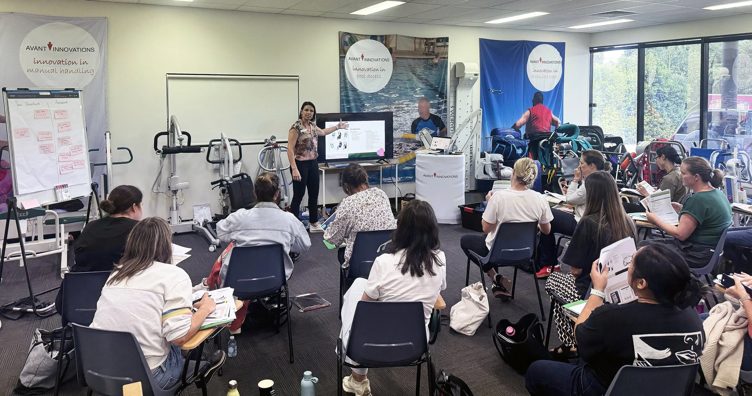A woman gives a presentation to an audience in a room filled with medical equipment and supplies, with banners displaying 'Avant Innovations' in the background.
