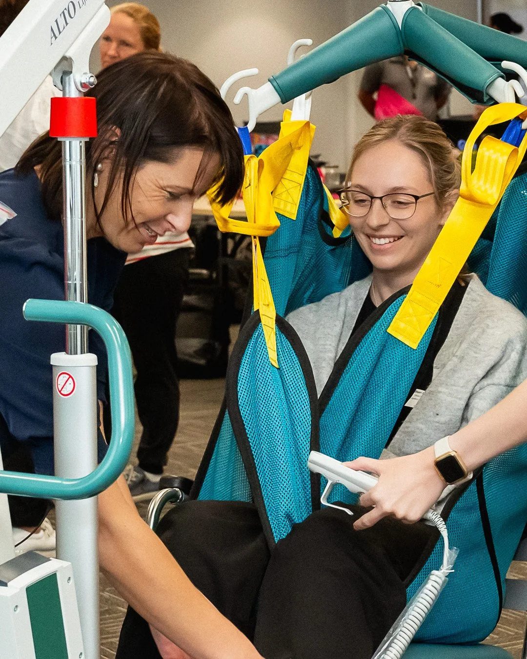 A woman smiling at a woman in a wheelchair, who is seated in a specialized therapy or diagnostic device with multicolored straps and harnesses. The woman in the wheelchair is wearing glasses and a gray shirt, and is holding a remote control. Another person is partially visible holding the remote. There are people and equipment in the background.