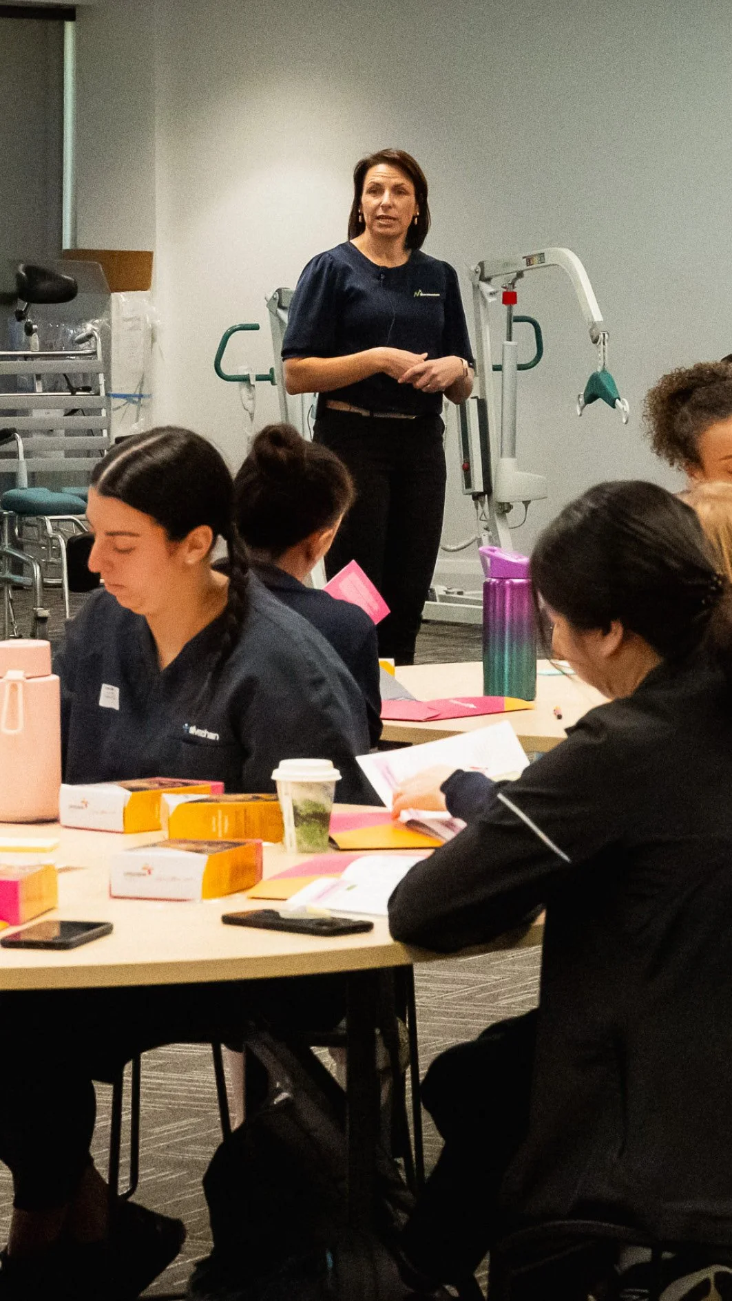 A woman stands at the front of a room, possibly giving a presentation or lecture. Several women are seated at tables, reading or writing, with notebooks, papers, and beverages in front of them. The background includes medical or exercise equipment.