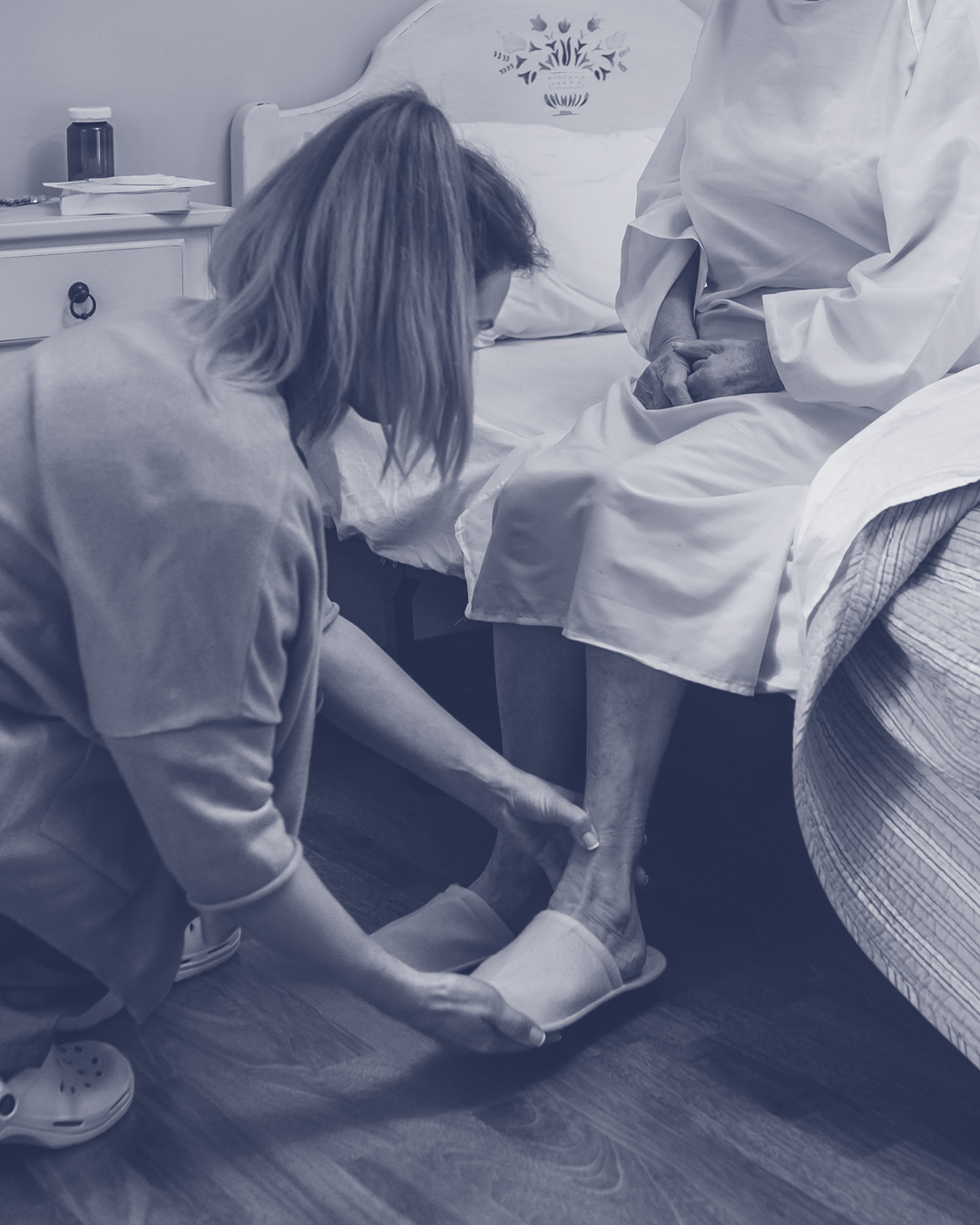 A woman helping an elderly person put on slippers in a bedroom.