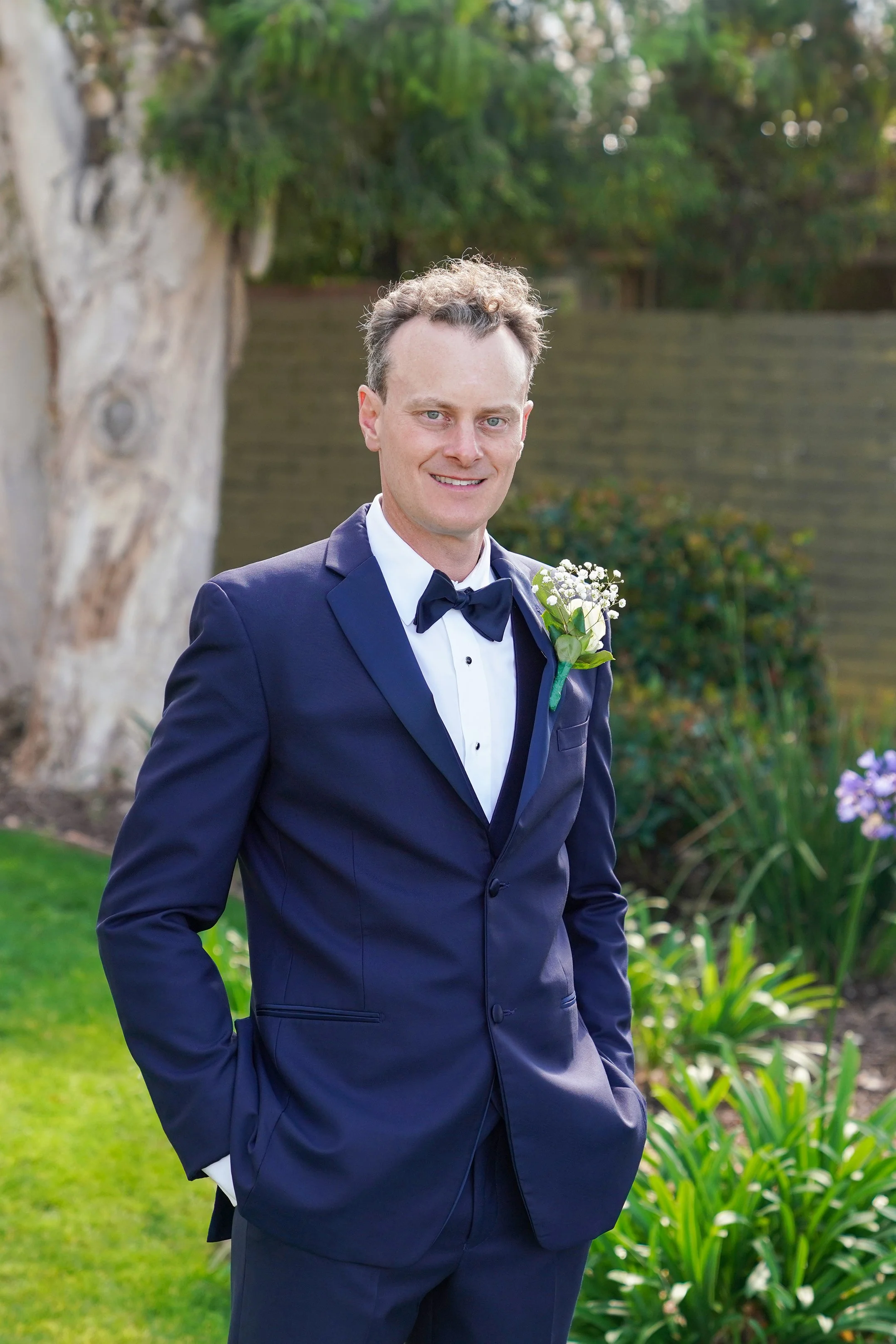 A man in a navy tuxedo with a bow tie and boutonnière stands outdoors in a garden with green plants and trees, smiling at the camera.