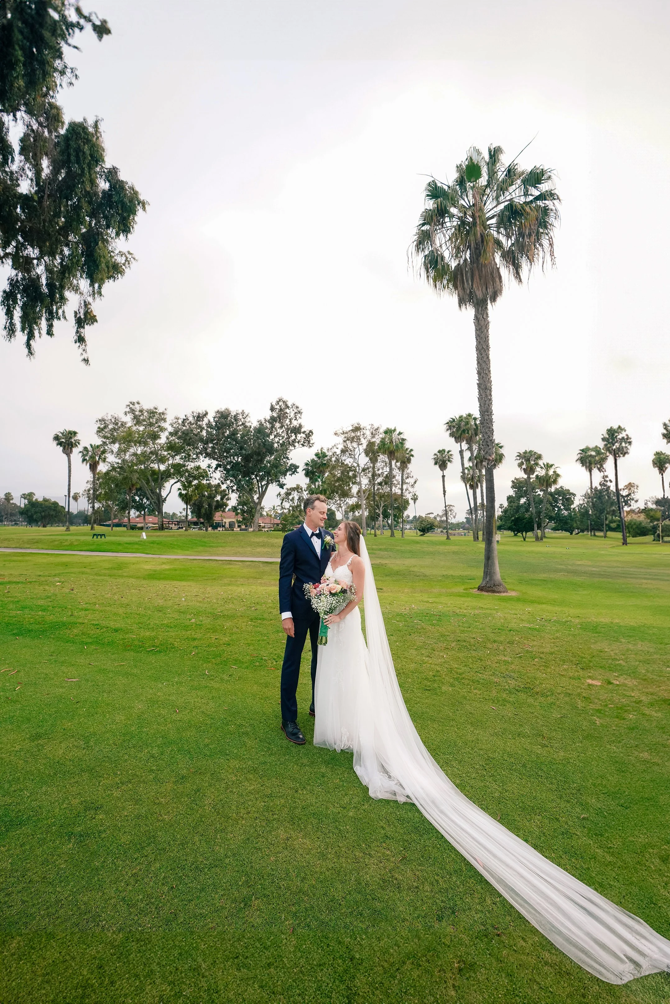 A bride and groom standing on a grassy field in a park, surrounded by tall trees and palm trees. The groom is in a navy tuxedo, and the bride is in a white wedding gown holding a bouquet. The bride's long veil trails on the ground behind her.