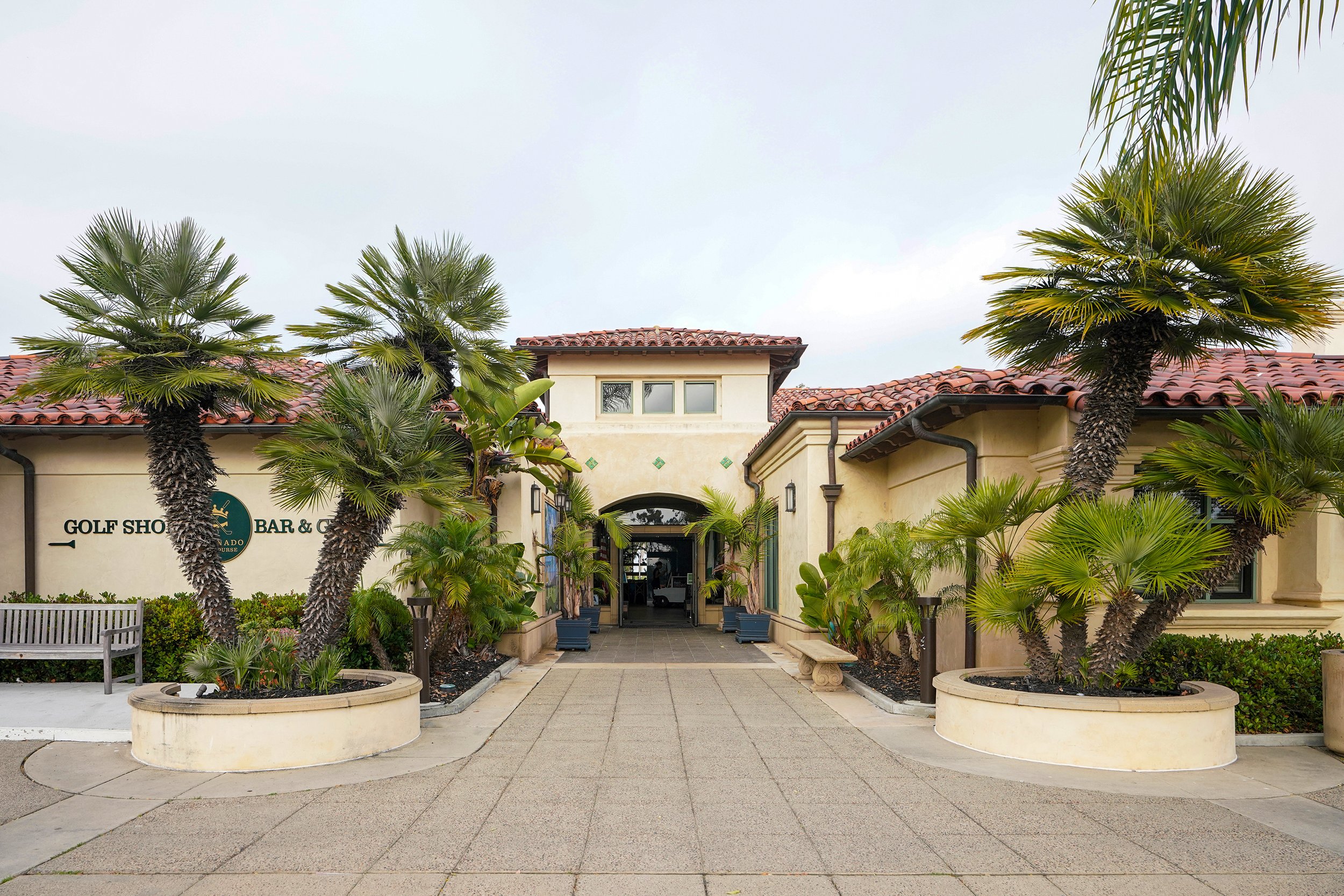 Entrance to a building with a sign reading 'Golf Shop, Bar & Grille,' framed by palm trees and lush plants on either side.
