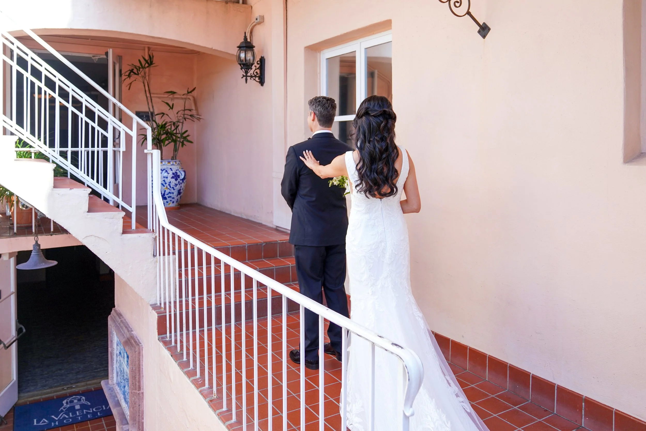 Bride in a V-neck lace wedding dress tapping the groom's shoulder during their first look portrait on an outdoor walkway at La Valencia Hotel in La Jolla.