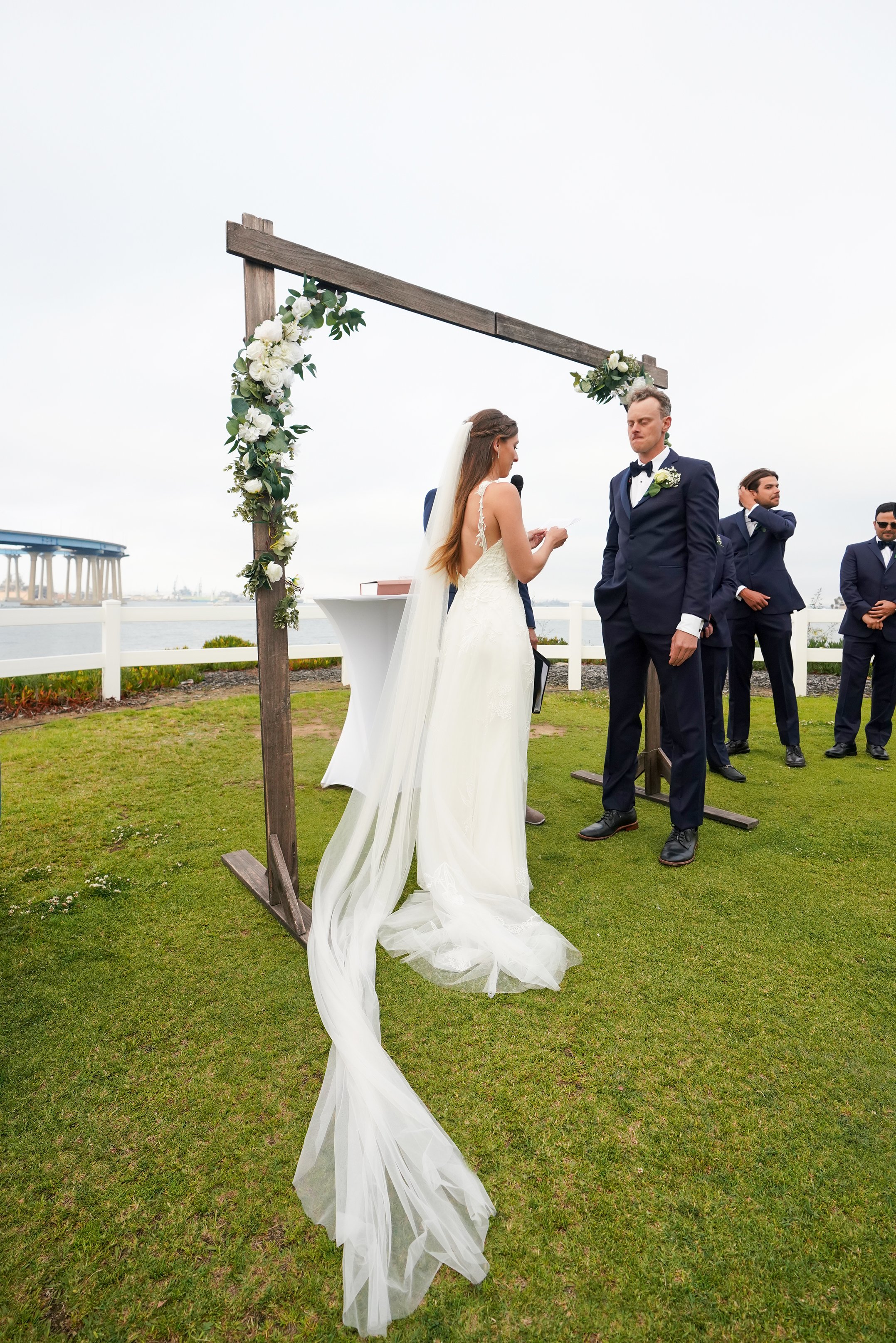 A full-length shot of a bride reading vows to her groom beneath a wooden arch decorated with white flowers. The bride wears a white gown with an ultra-long veil puddling on the grass. The groom and his groomsmen wear navy blue suits. The San Diego-Co