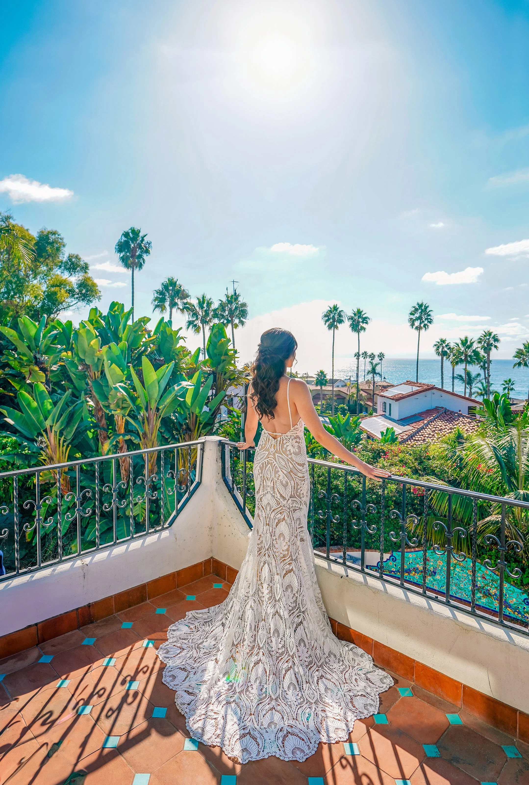 A bride from behind stands on a tiled balcony looking out at the ocean on a sunny day. She wears a backless lace wedding gown with spaghetti straps and a detailed train. The coastal view features palm trees, tropical greenery.