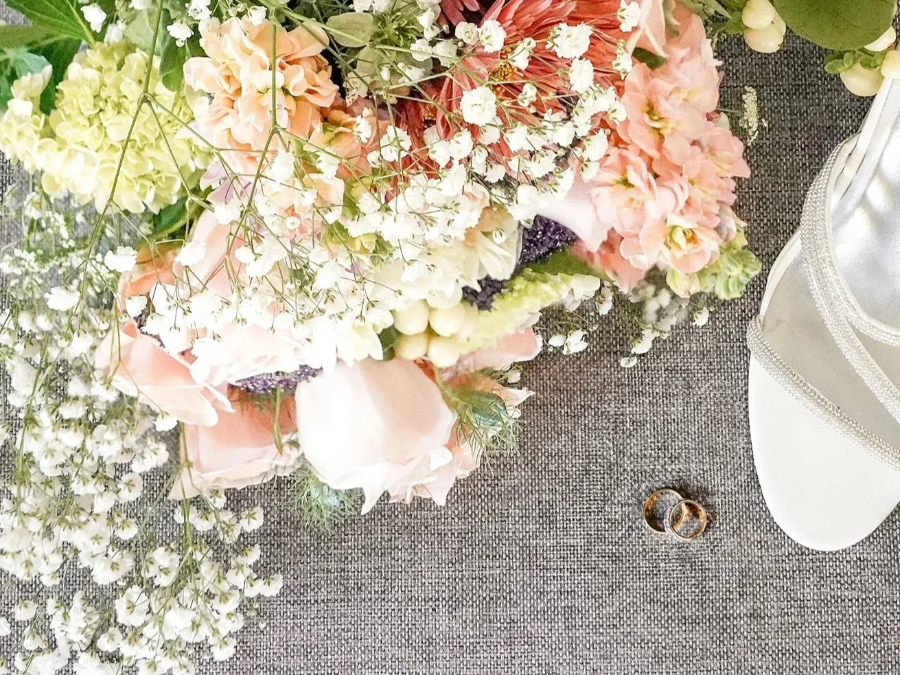 A large floral arrangement with pink, white, and purple flowers on a table, next to a white decorative stand and two wedding rings.