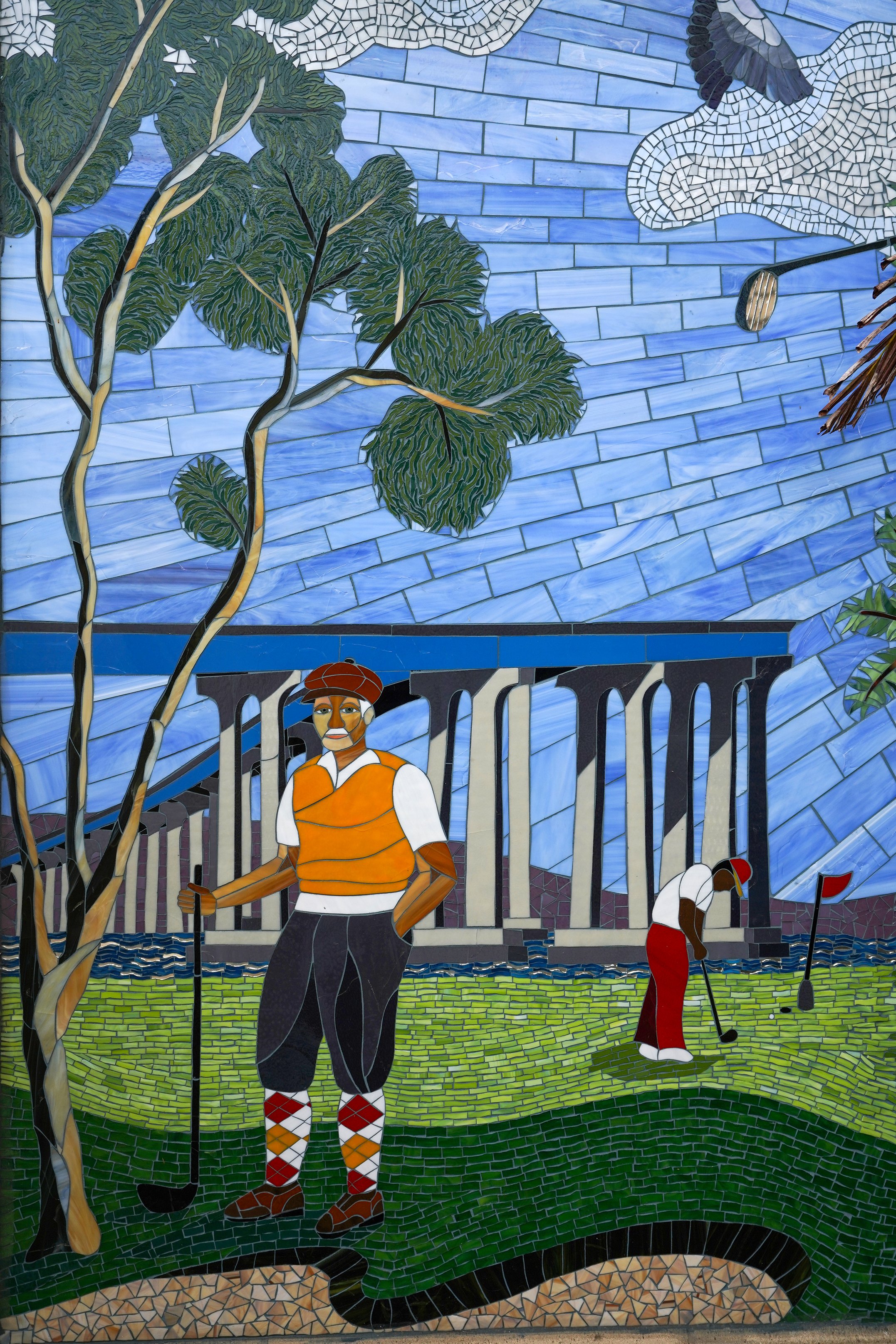 Colorful mosaic mural depicting a golfer in a red and white outfit preparing to tee off on a golf course, with trees, a blue sky, and buildings in the background.