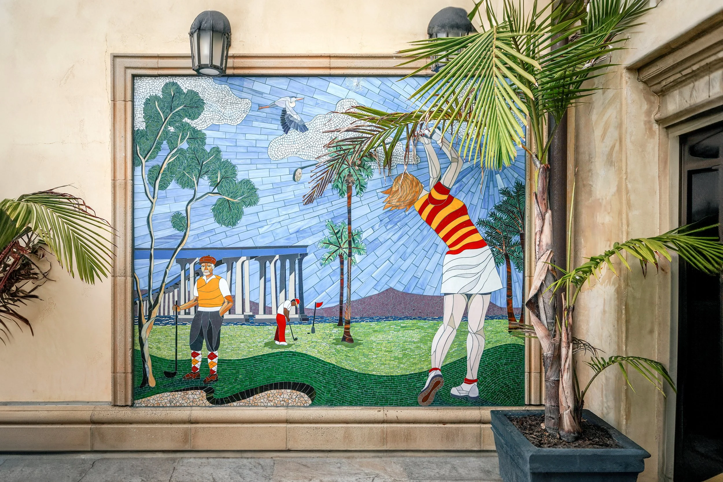 A colorful outdoor mosaic mural depicting golfers, a woman, trees, clouds, and a cypress tree with a plant in a blue pot in the foreground.
