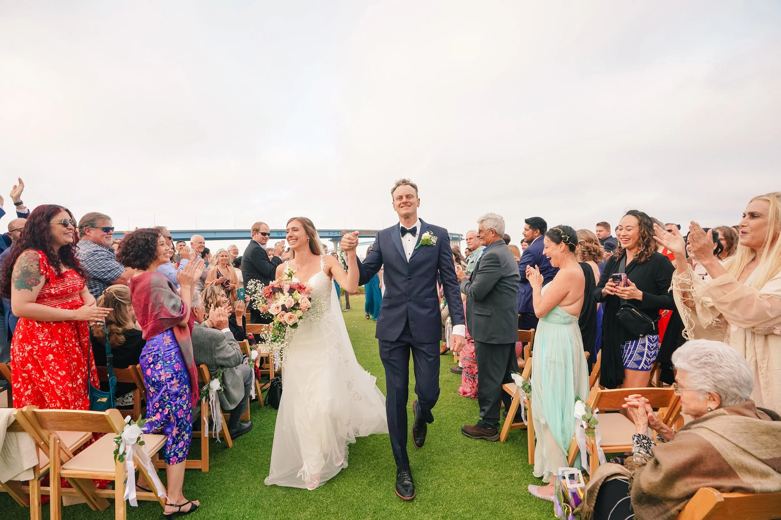 A wide, horizontal shot of a newly married bride and groom smiling and holding hands while walking down the aisle at an outdoor wedding ceremony. Cheering guests line both sides of the grass aisle in folding wooden chairs. The blue San Diego-Coronado