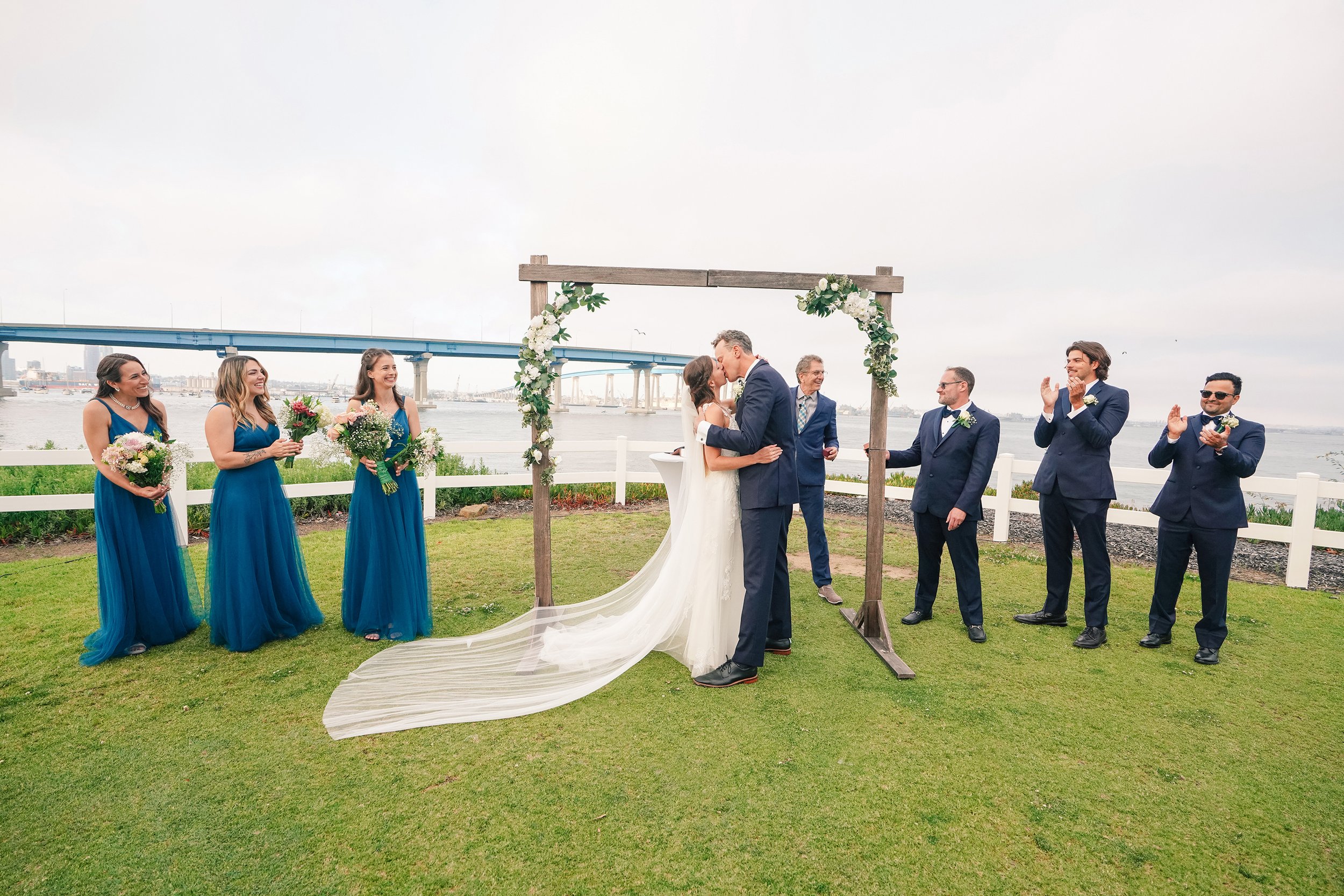 A wedding ceremony taking place outdoors by the water with a bride and groom kissing under a floral-covered wooden arch while bridesmaids and groomsmen stand on either side, clapping and smiling.