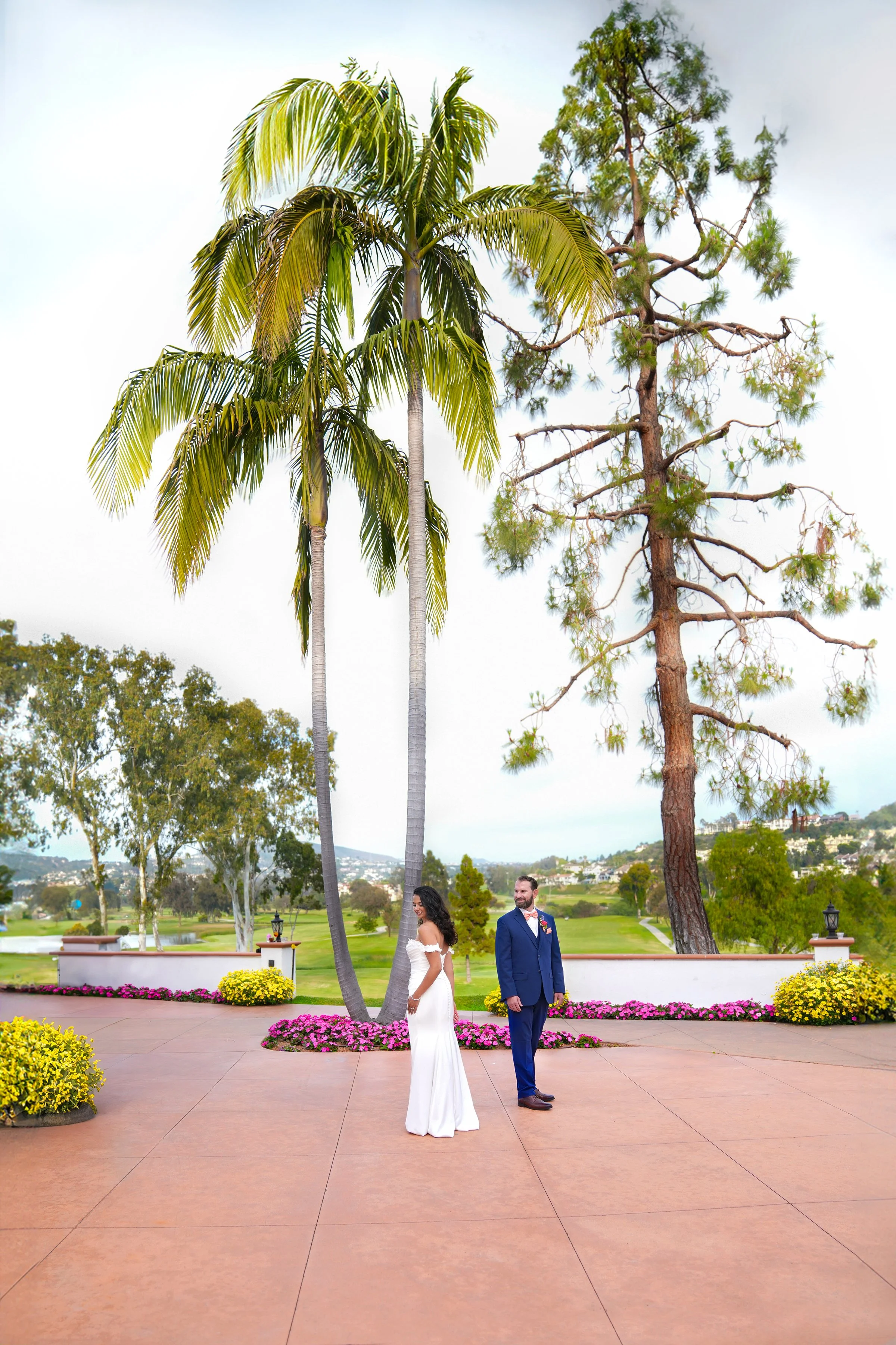 Bride in an off-the-shoulder white dress and groom in a blue suit standing outdoors on a patio between a palm tree and a pine tree.