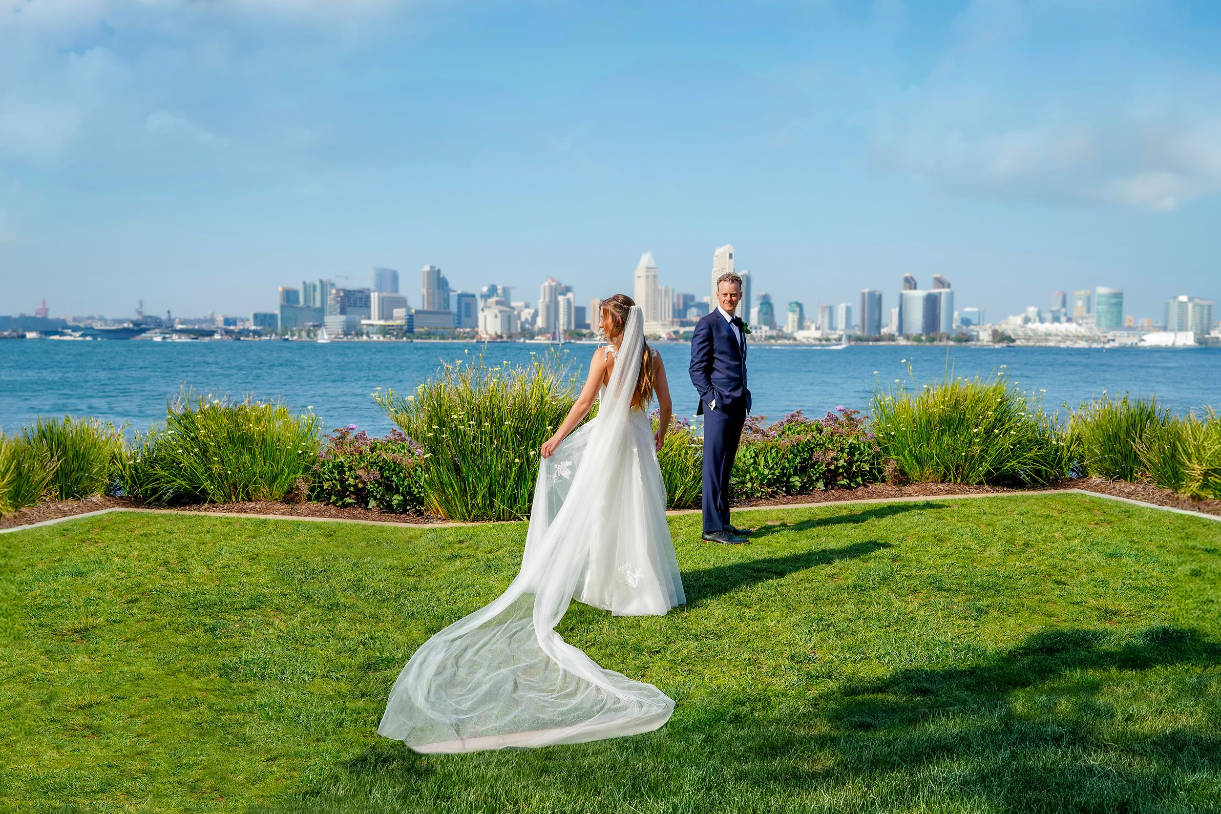 A bride in an elegant wedding gown with a long flowing veil and a groom in a navy suit posing on a grassy lawn with the downtown San Diego skyline and bay in the background on Coronado Island.