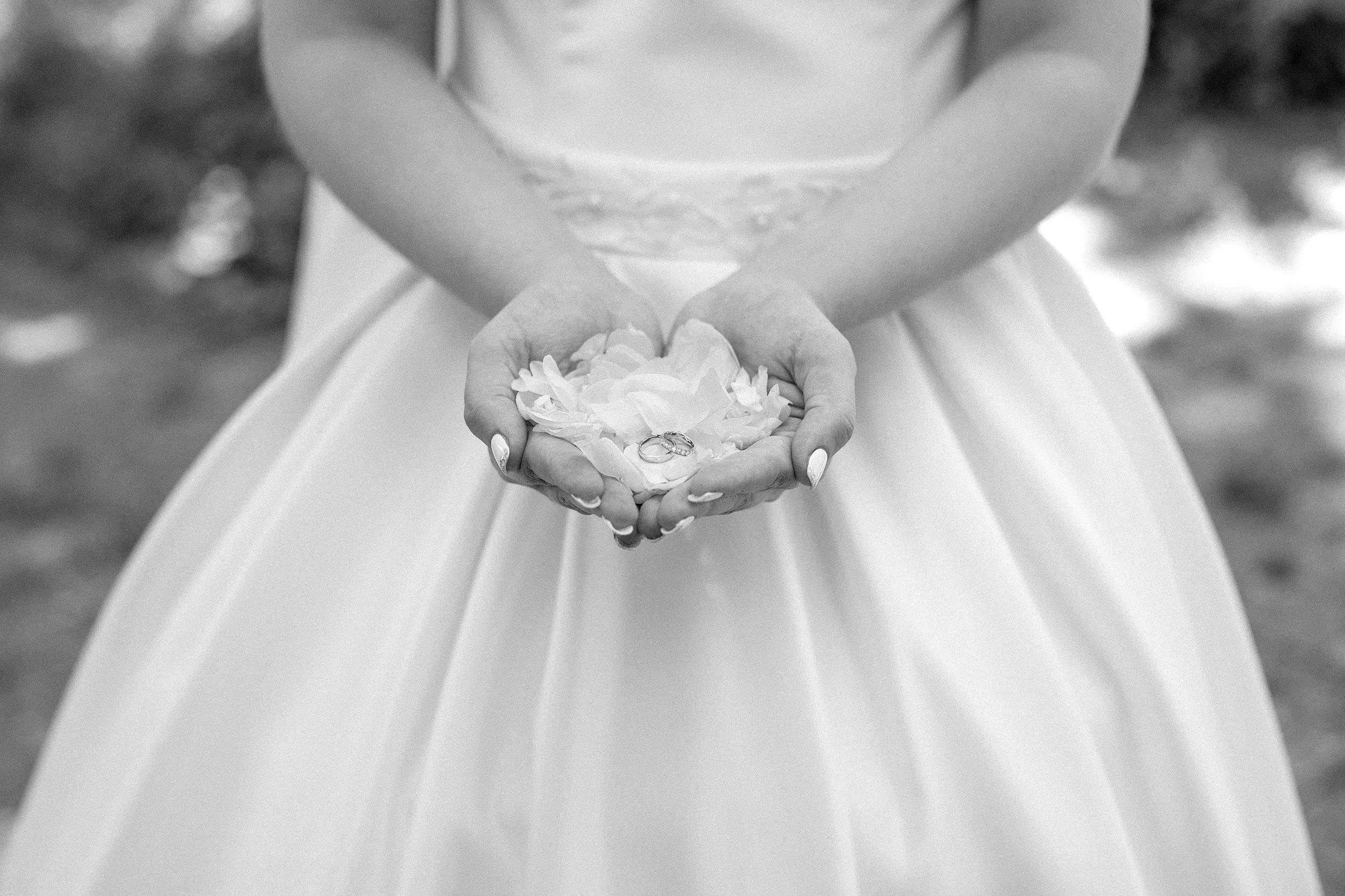 An elegant, black and white close-up photo focusing on a bride’s cupped hands holding a small nest of white flowers. Two gold wedding rings sit nestled in the center of the petals. The bride wears a classic, textured white wedding dress that fades.