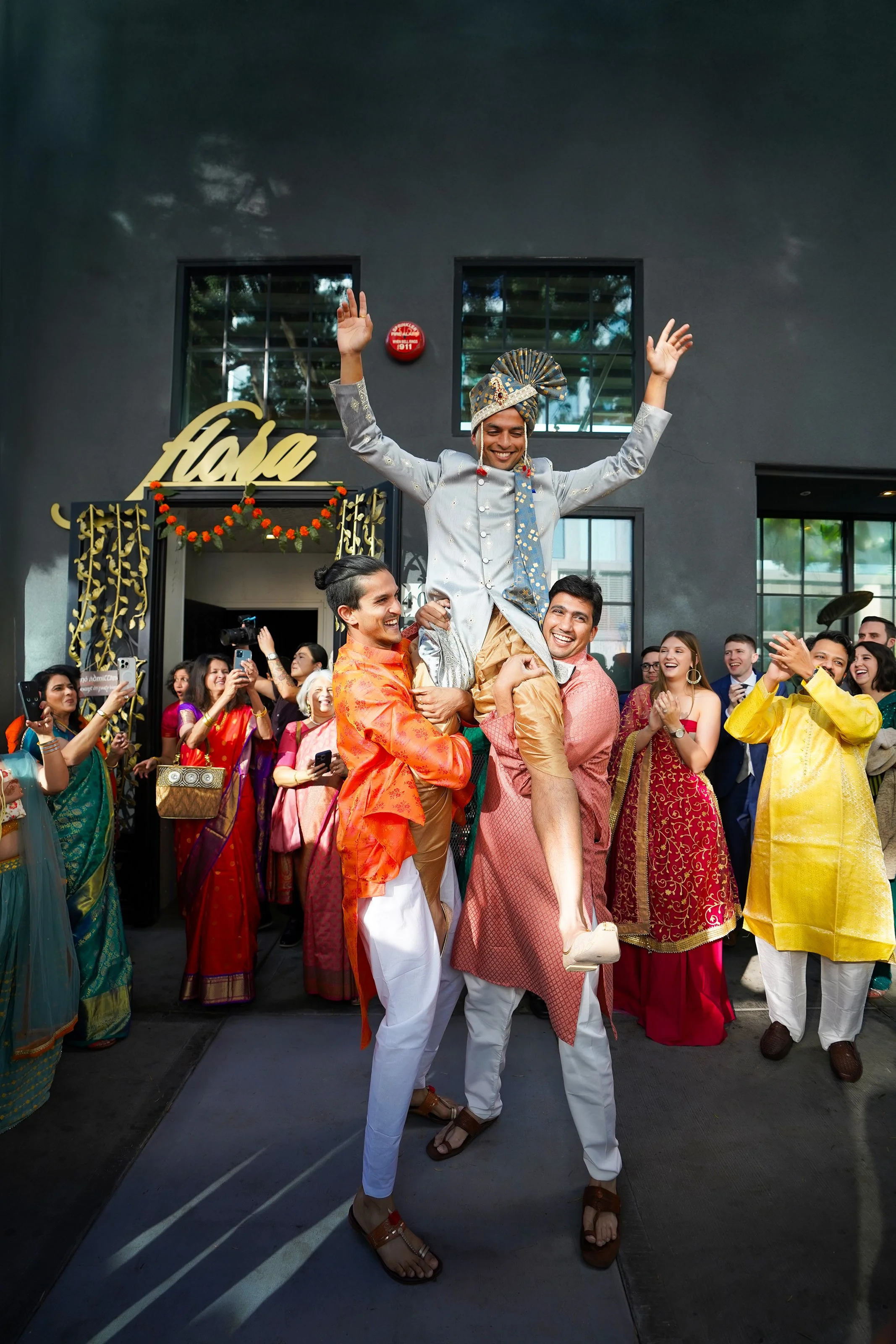 A group of people joyfully celebrates an Indian wedding outside Flora wedding venue. Two men carry the groom on their shoulders as he lifts both arms in excitement.