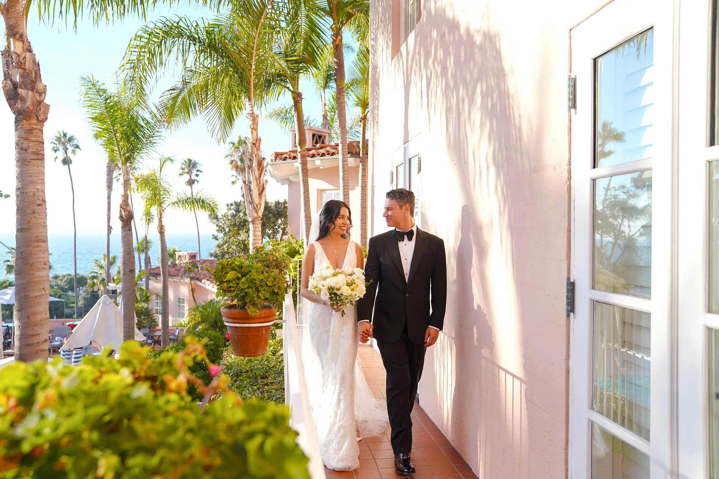 A bright, romantic wedding portrait of a bride and groom holding hands while walking along an outdoor terracotta balcony at La Valencia Hotel in La Jolla, San Diego. The bride wears a plunging V-neck lace wedding dress with a long bridal veil and hol
