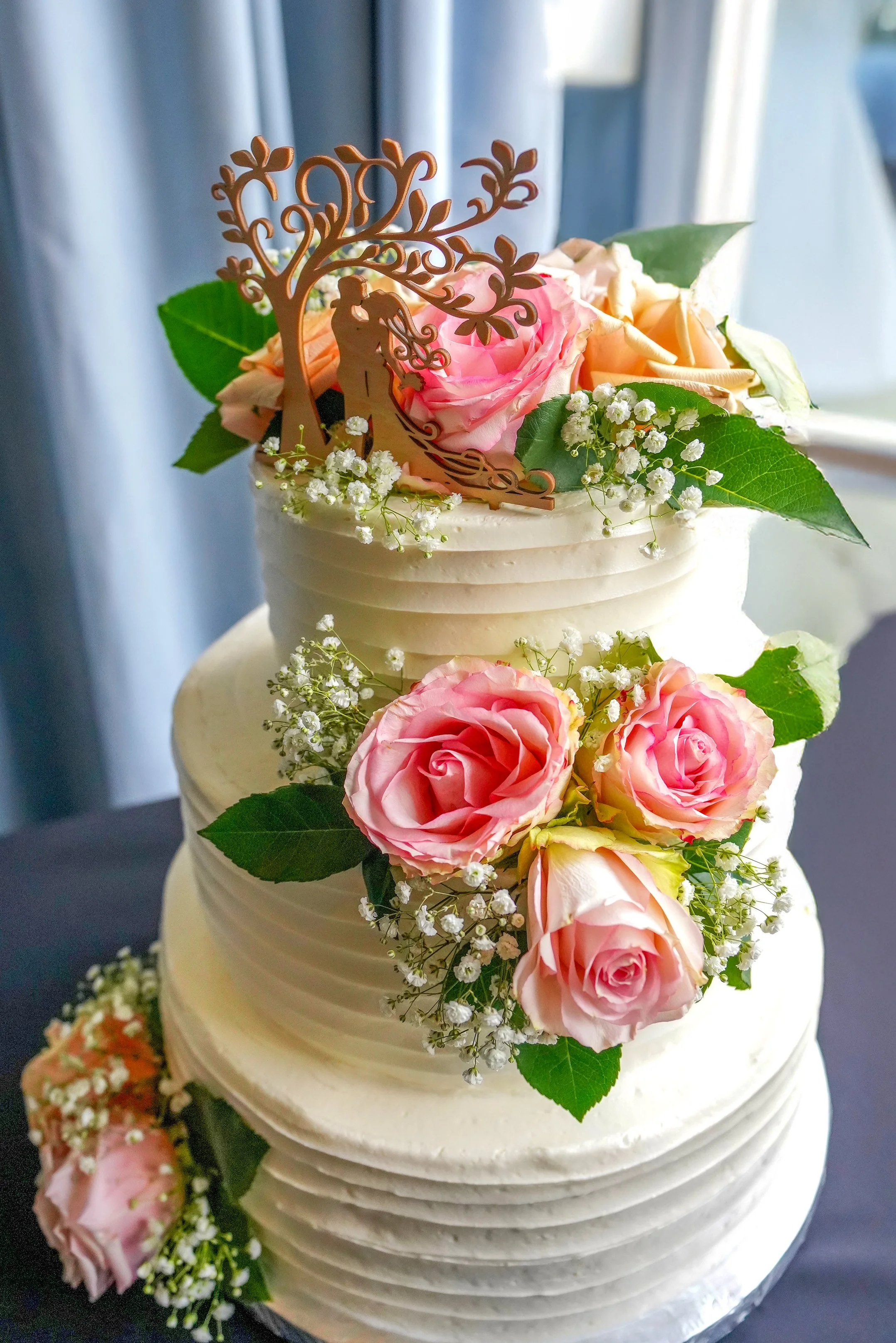 A multi-layered white wedding cake decorated with pink and peach roses, green leaves, baby's breath flowers, and a wooden cake topper of a couple under a tree.
