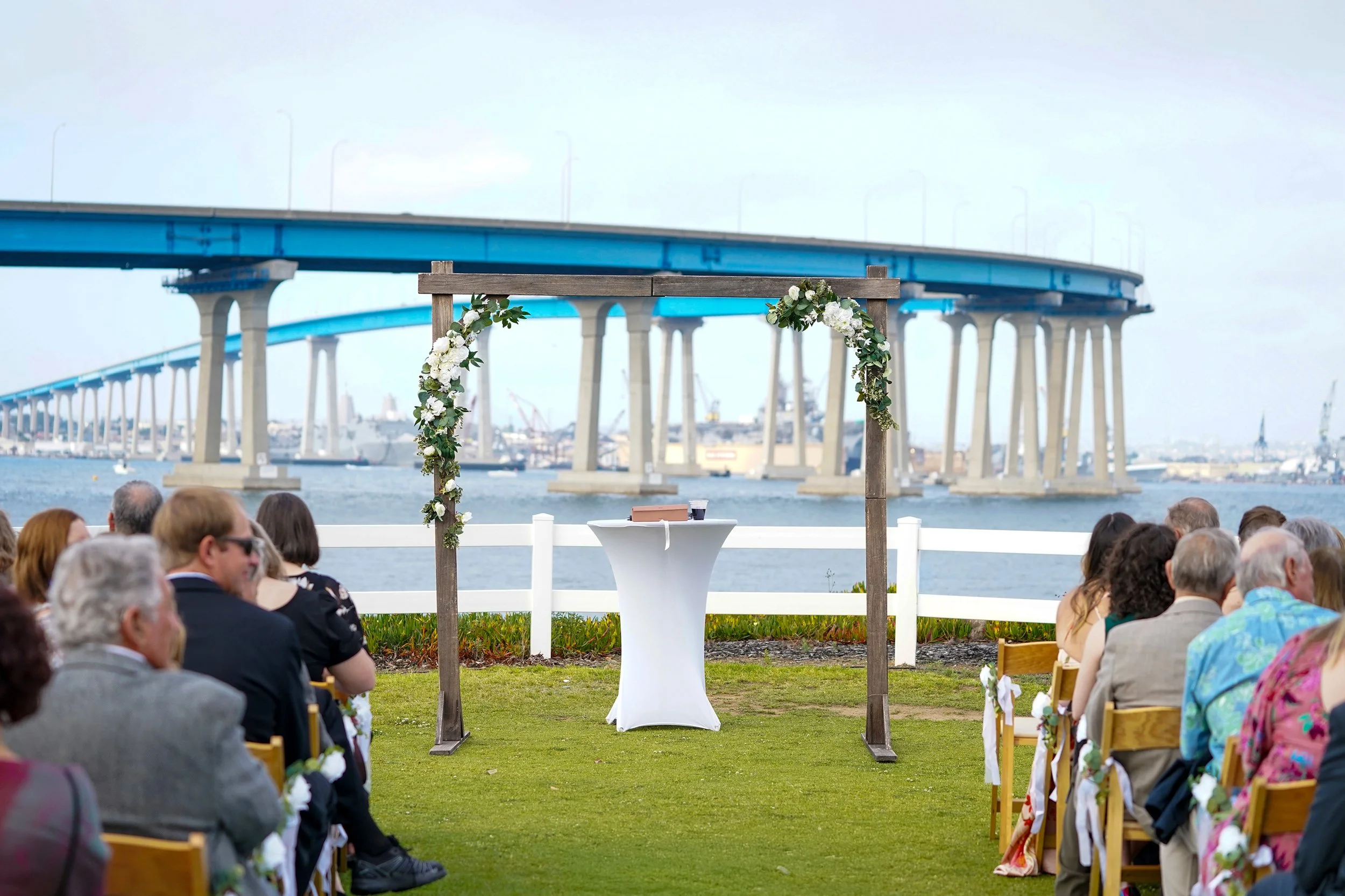 Outdoor wedding ceremony area near a bridge, with seated guests on both sides and a white table under a floral arch.