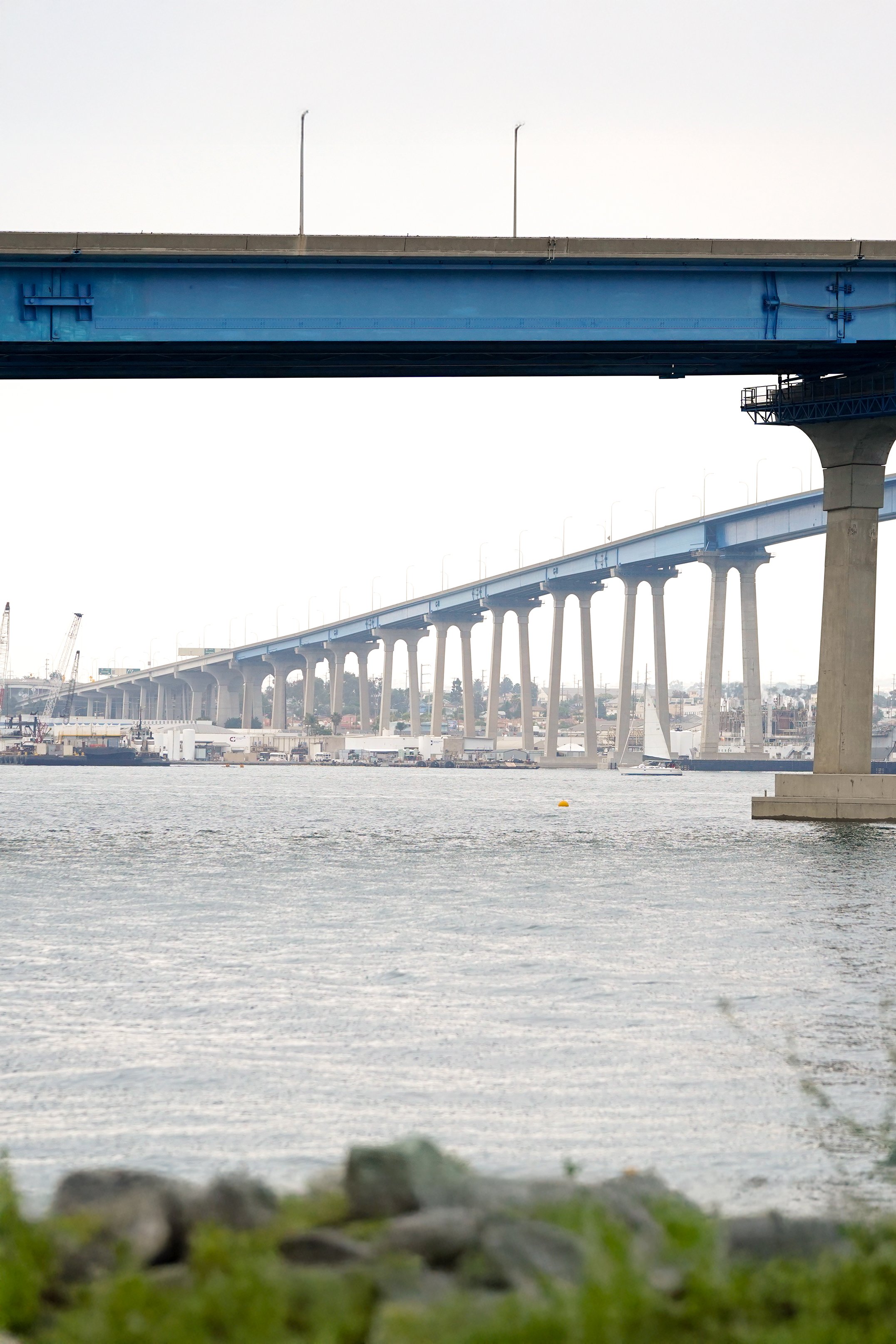 View of a cityscape with a large, elevated blue bridge over a body of water. Industrial buildings and cranes are visible in the background, with some boats on the water and greenery in the foreground.