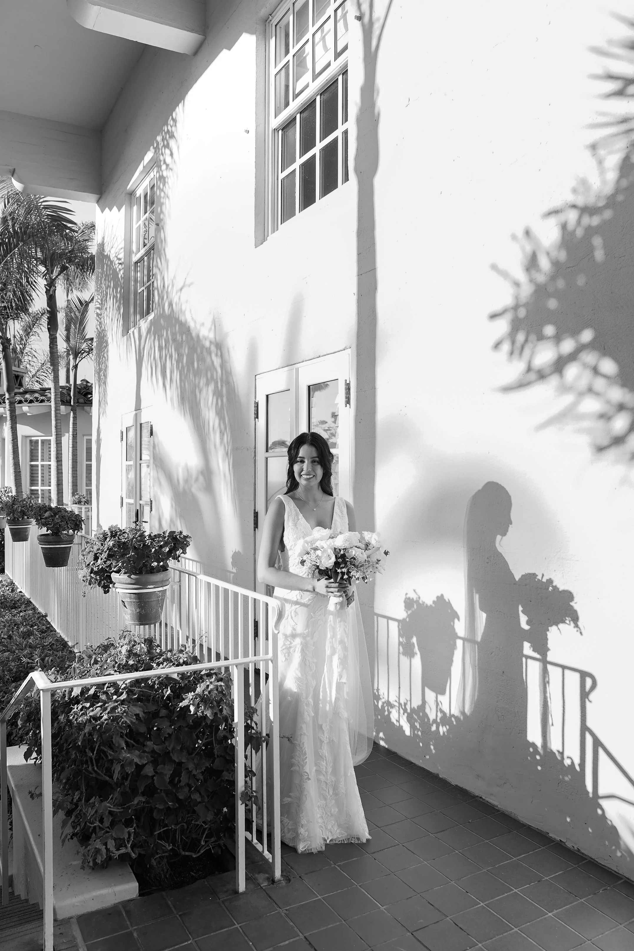 Black and white bridal portrait of a smiling bride holding a white floral bouquet on an outdoor patio walkway at La Valencia Hotel in La Jolla.