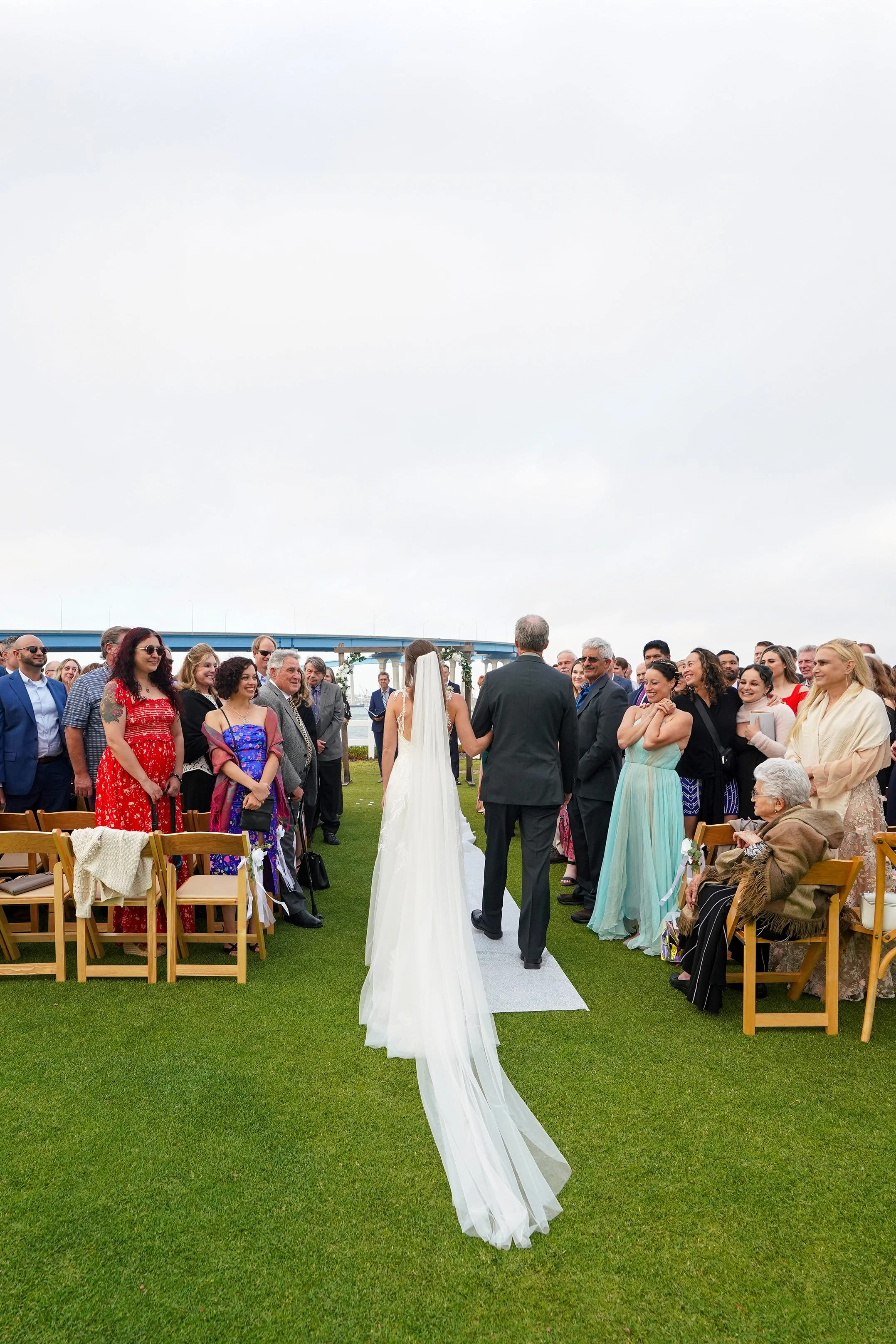 A wedding ceremony outdoors with a bride and groom holding hands, surrounded by guests seated on wooden chairs on a grassy area under a cloudy sky.