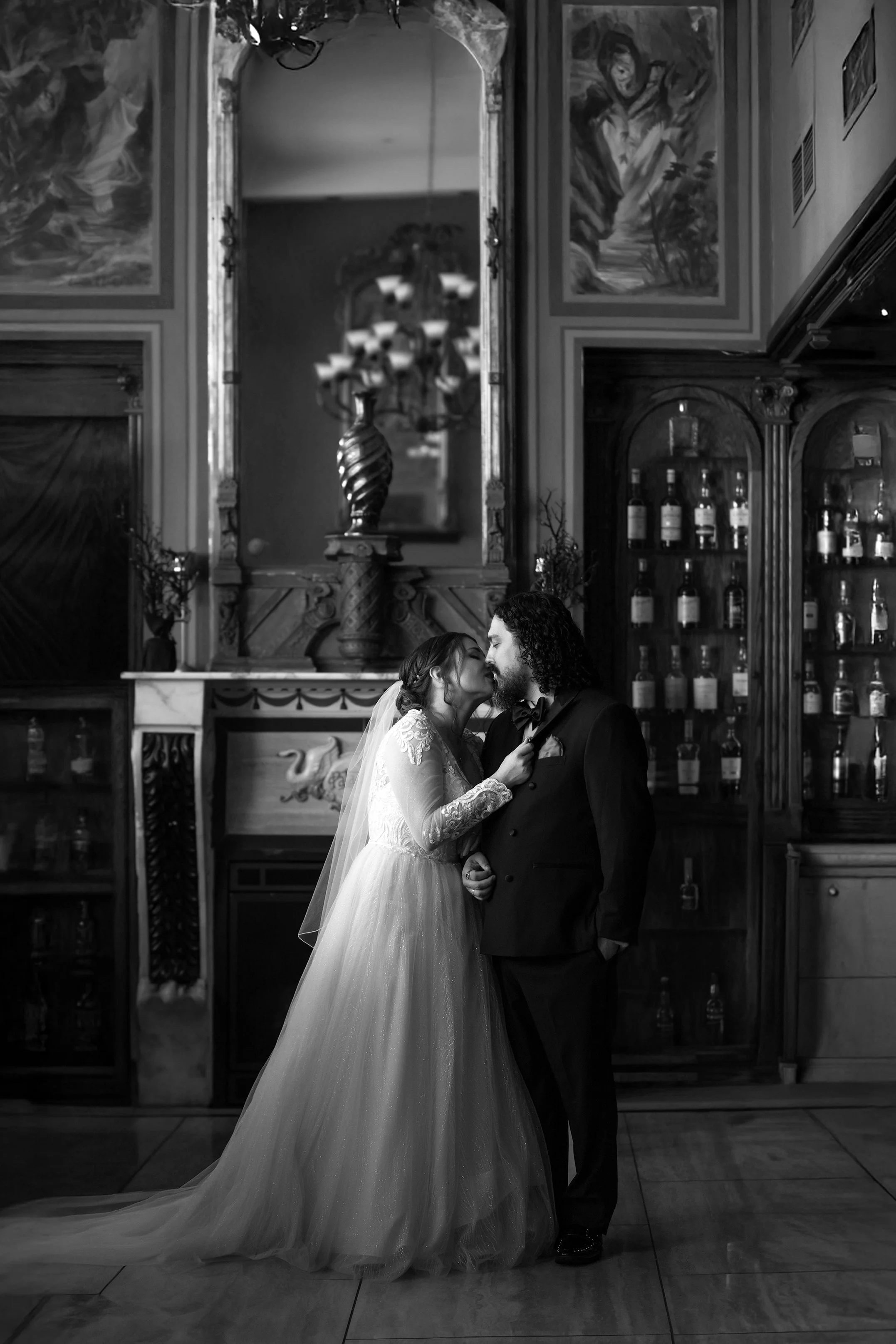 Black and white wedding portrait of a bride in a long-sleeve lace gown and veil kissing her groom in front of a moody, ornate vintage bar and fireplace.