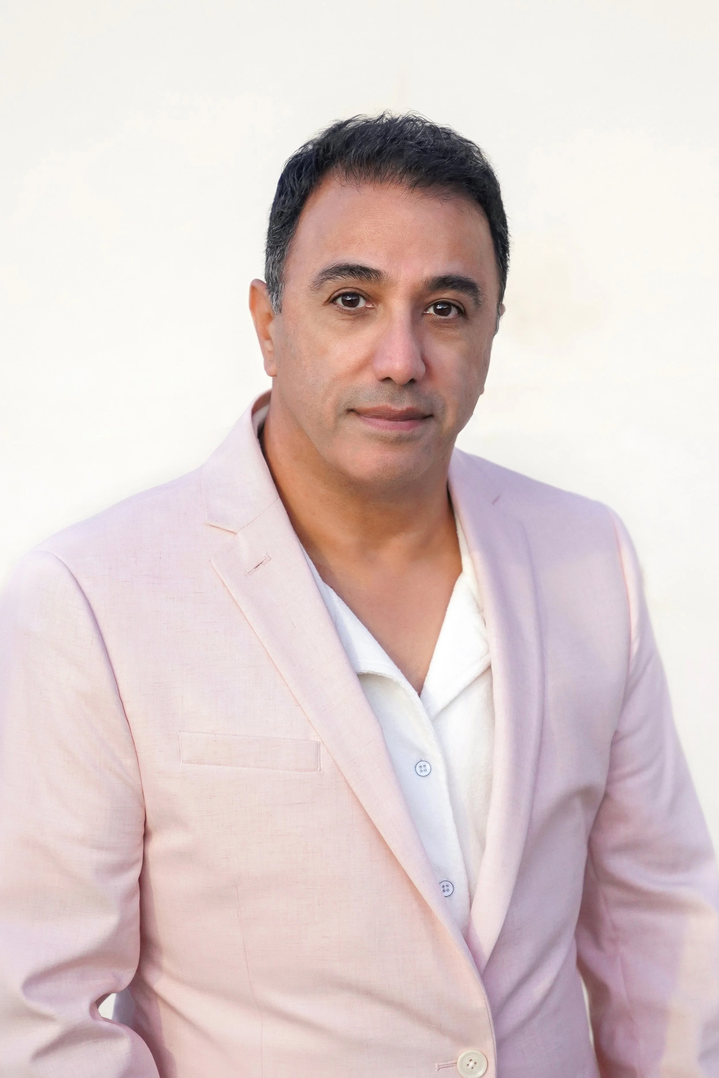 A man with dark hair wearing a light pink suit and white shirt, facing forward with a slight smile, against a plain white background.