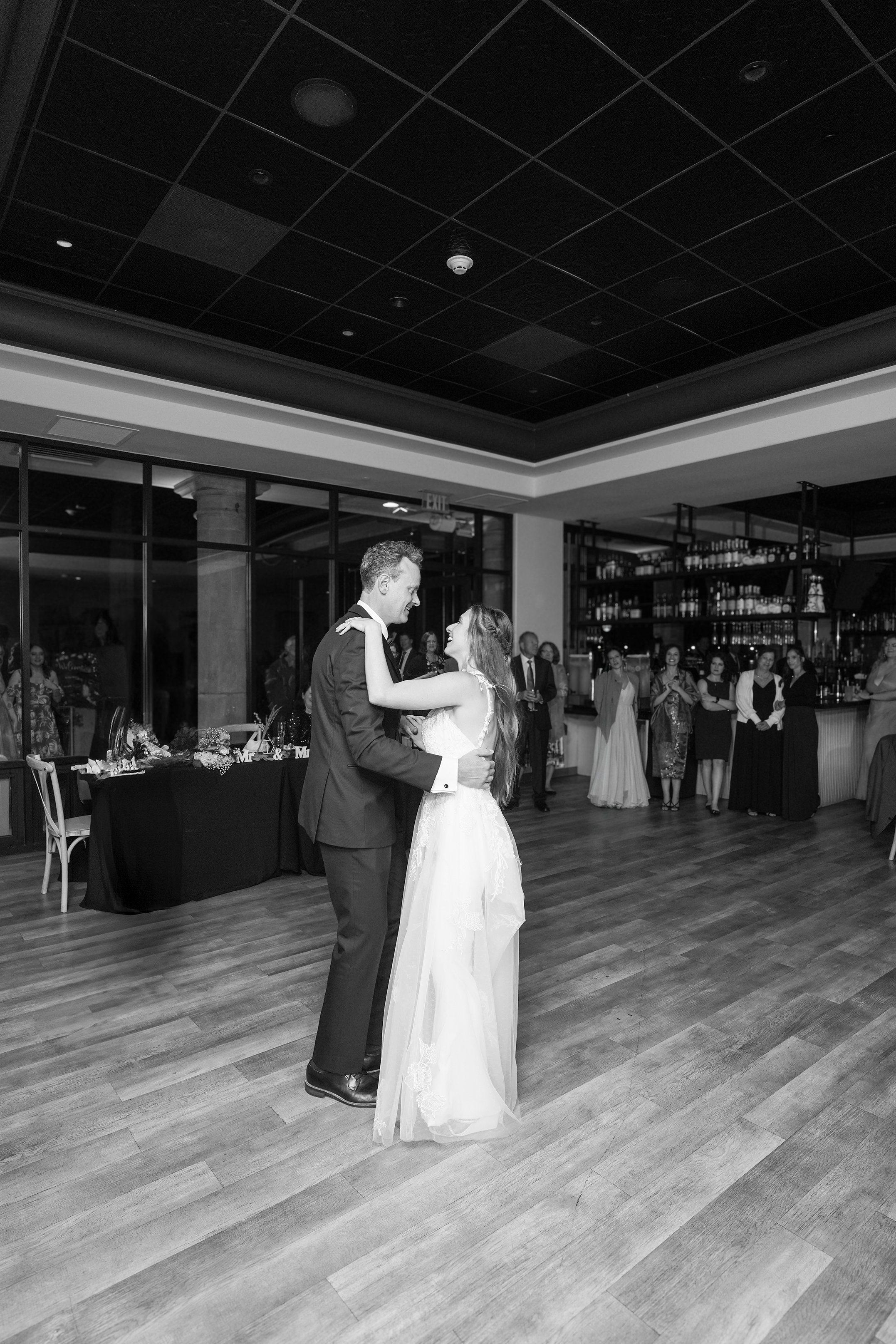 A bride and groom are dancing in a reception hall surrounded by guests, with a bar in the background and a table with flowers and decorations to the side.