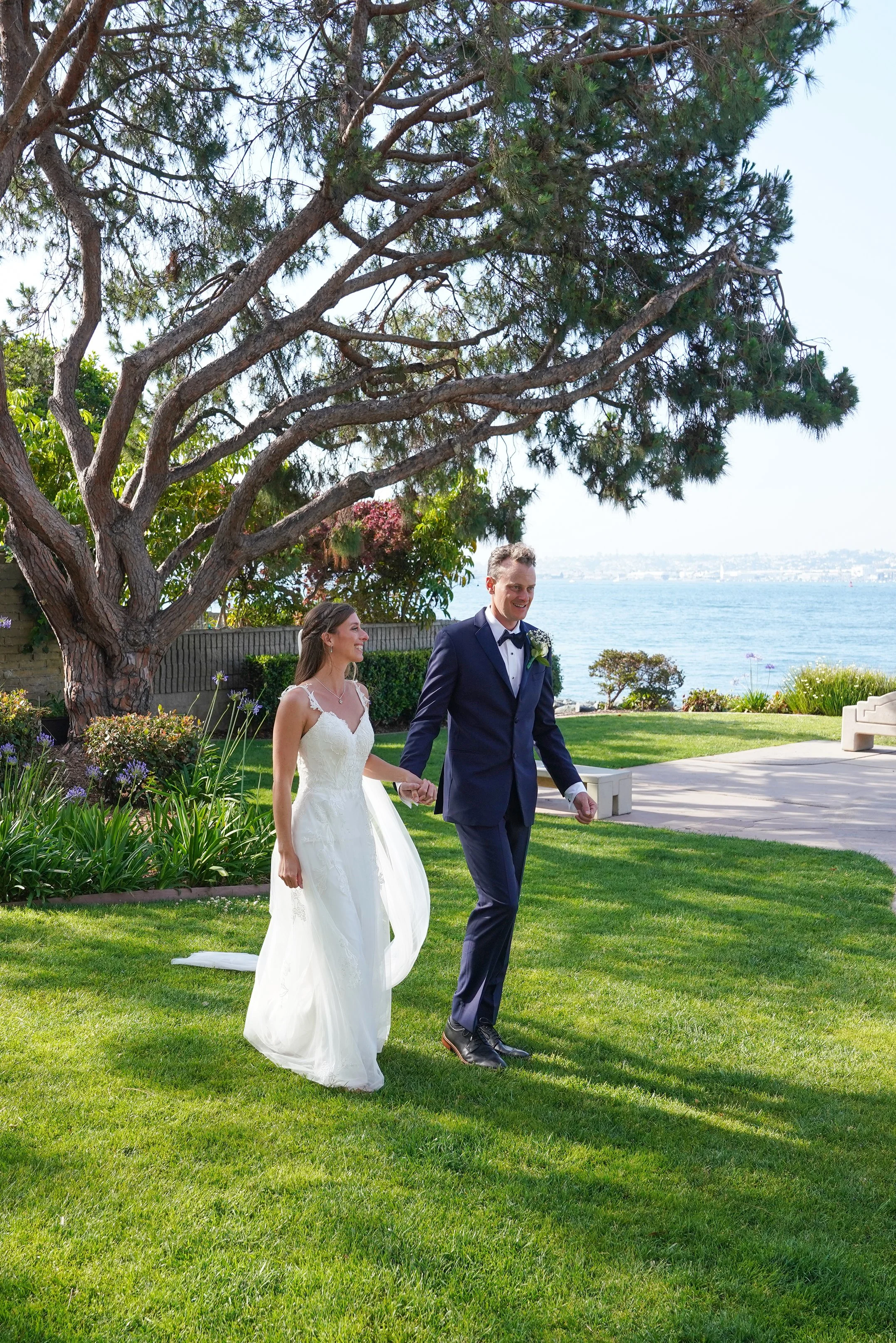 A bride and groom holding hands and walking on a grassy lawn near a body of water, with trees and distant shoreline in the background on a sunny day.