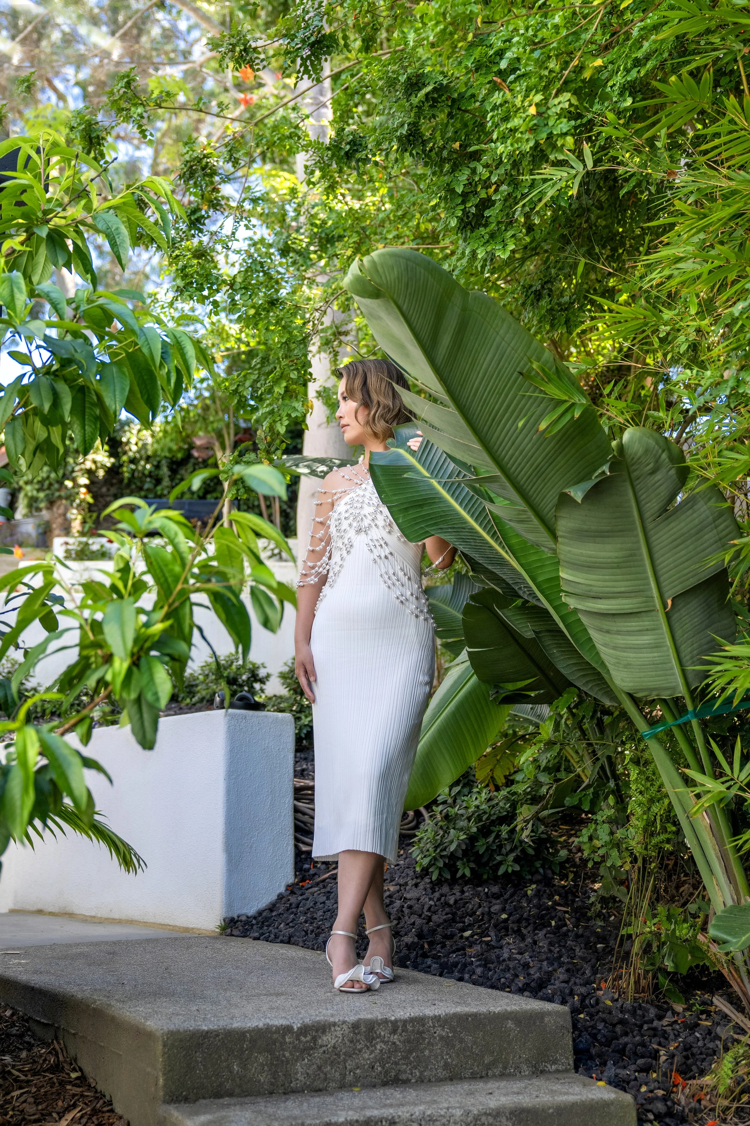Bride in a pleated white midi dress with a decorative pearl shoulder cape posing outdoors surrounded by lush tropical greenery.