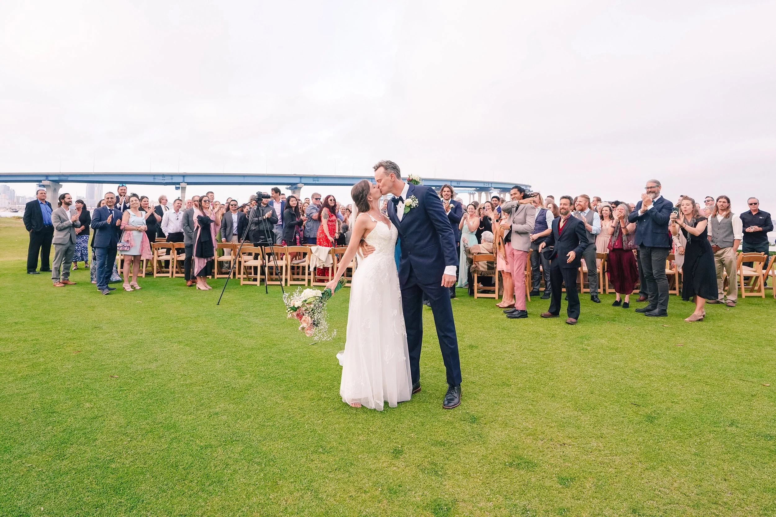 A bride and groom kissing at an outdoor wedding ceremony, with guests clapping and smiling in the background.