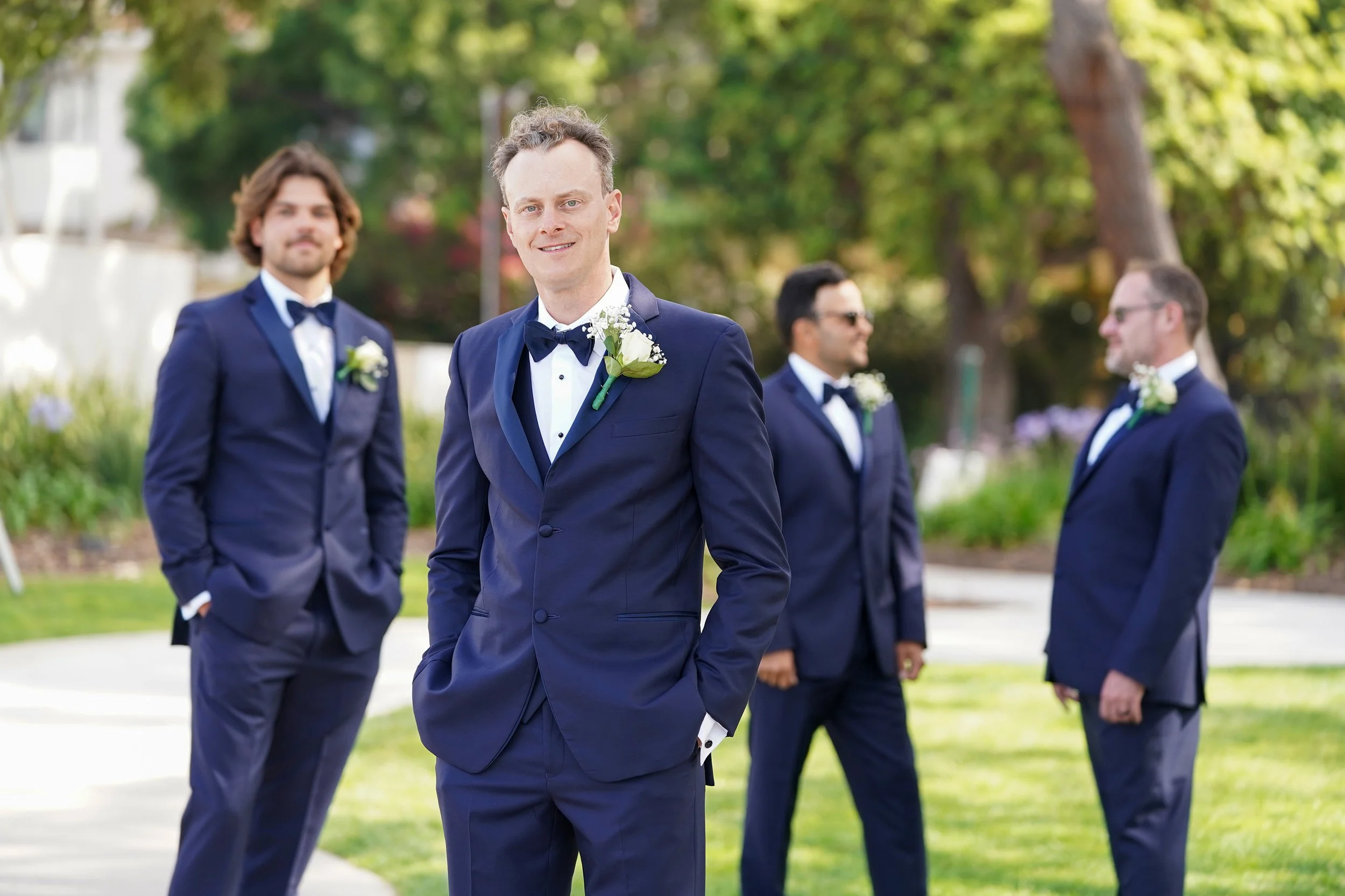 A group of men in navy blue tuxedos with white shirts and black bow ties, standing outdoors on a sunny day, with the man in the foreground smiling at the camera and other men in the background engaged in conversation.