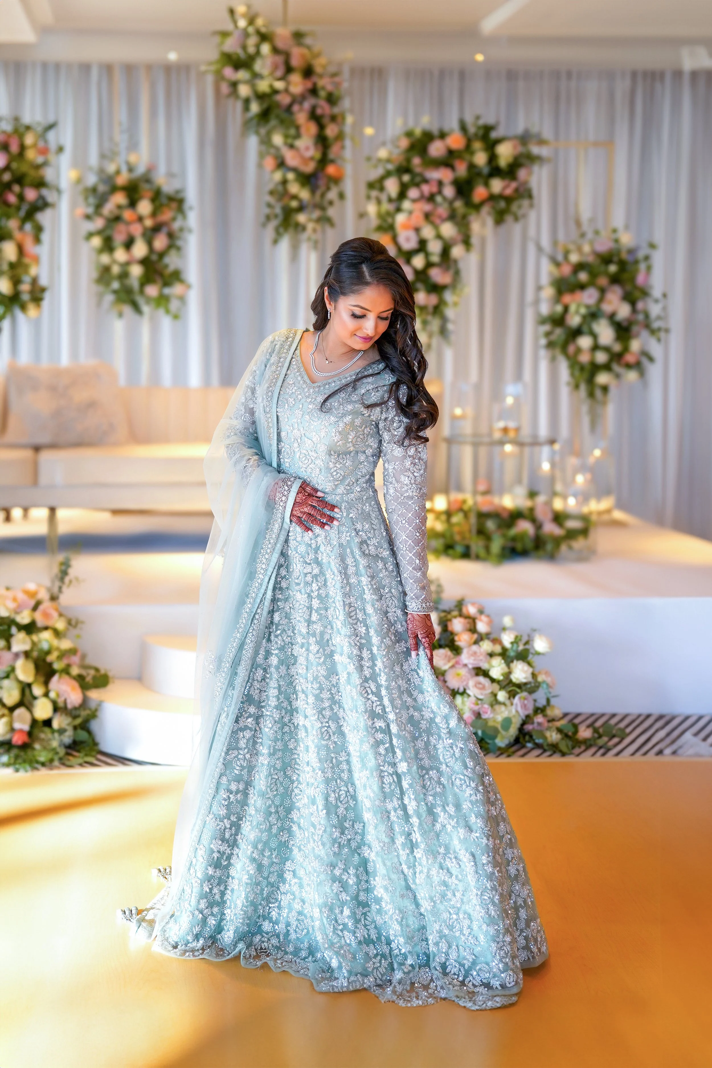 Elegant South Asian bride in an embroidered mint green anarkali gown posing at a luxury Indian wedding reception venue with pastel floral decorations.
