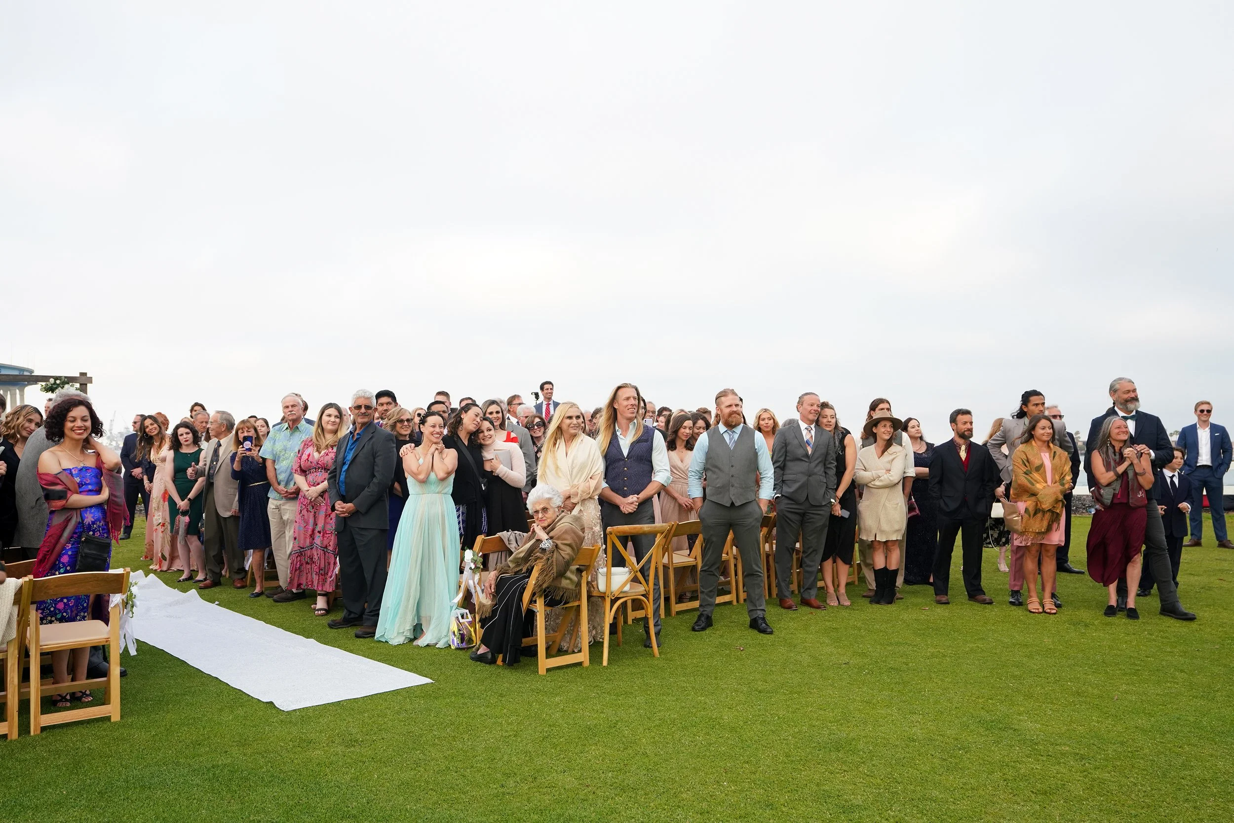 A large group of diverse people dressed in formal attire, gathered outdoors for a wedding ceremony, standing on a green lawn under an overcast sky.