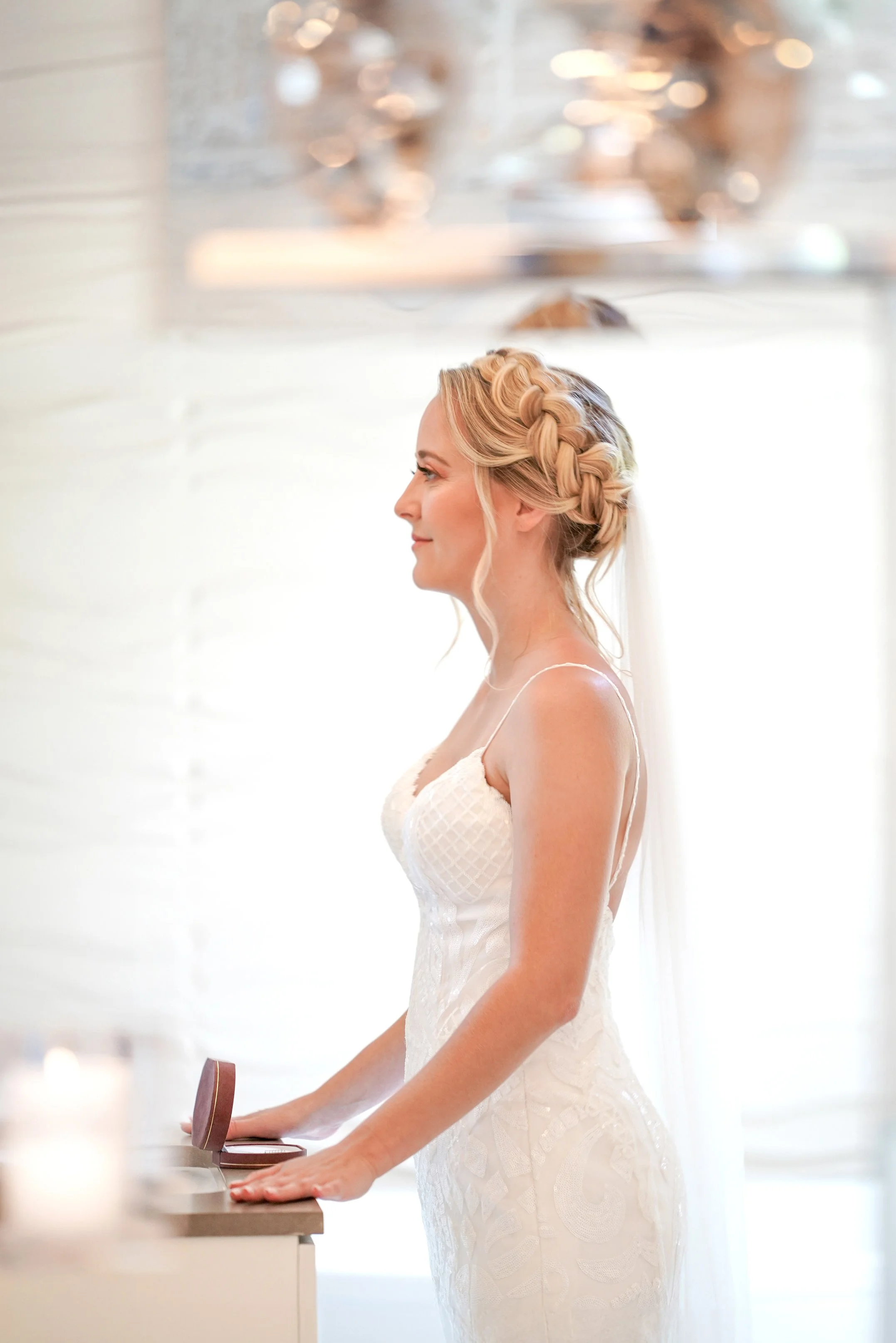 Side profile portrait of a blonde bride with an elegant braided updo hairstyle and long veil, wearing a textured white wedding dress.