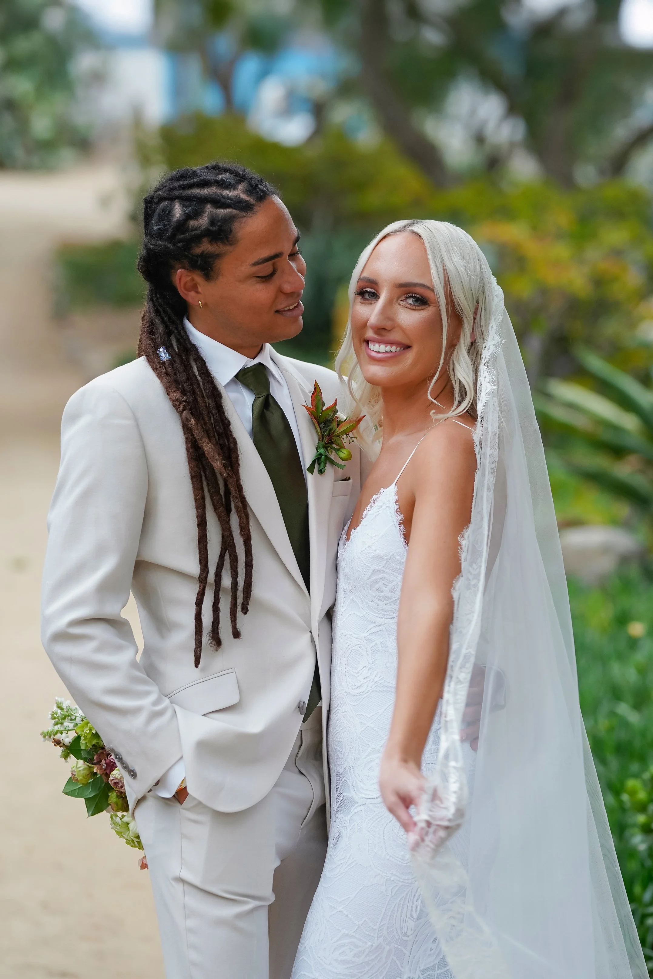 A wedding couple outdoors, the groom with long dreadlocks in a white suit and green tie, and the bride with platinum blonde hair in a white lace wedding dress and veil, smiling.