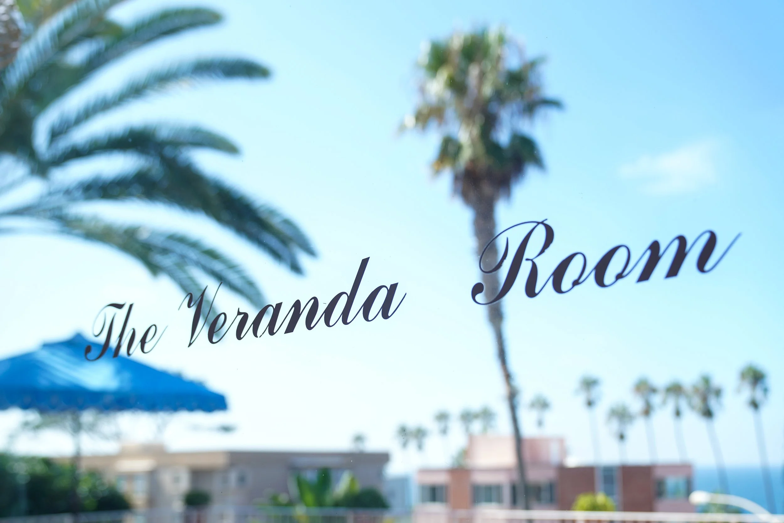 The Veranda Room sign on a glass window at La Valencia Hotel in La Jolla with blurred palm trees and a blue sky background.