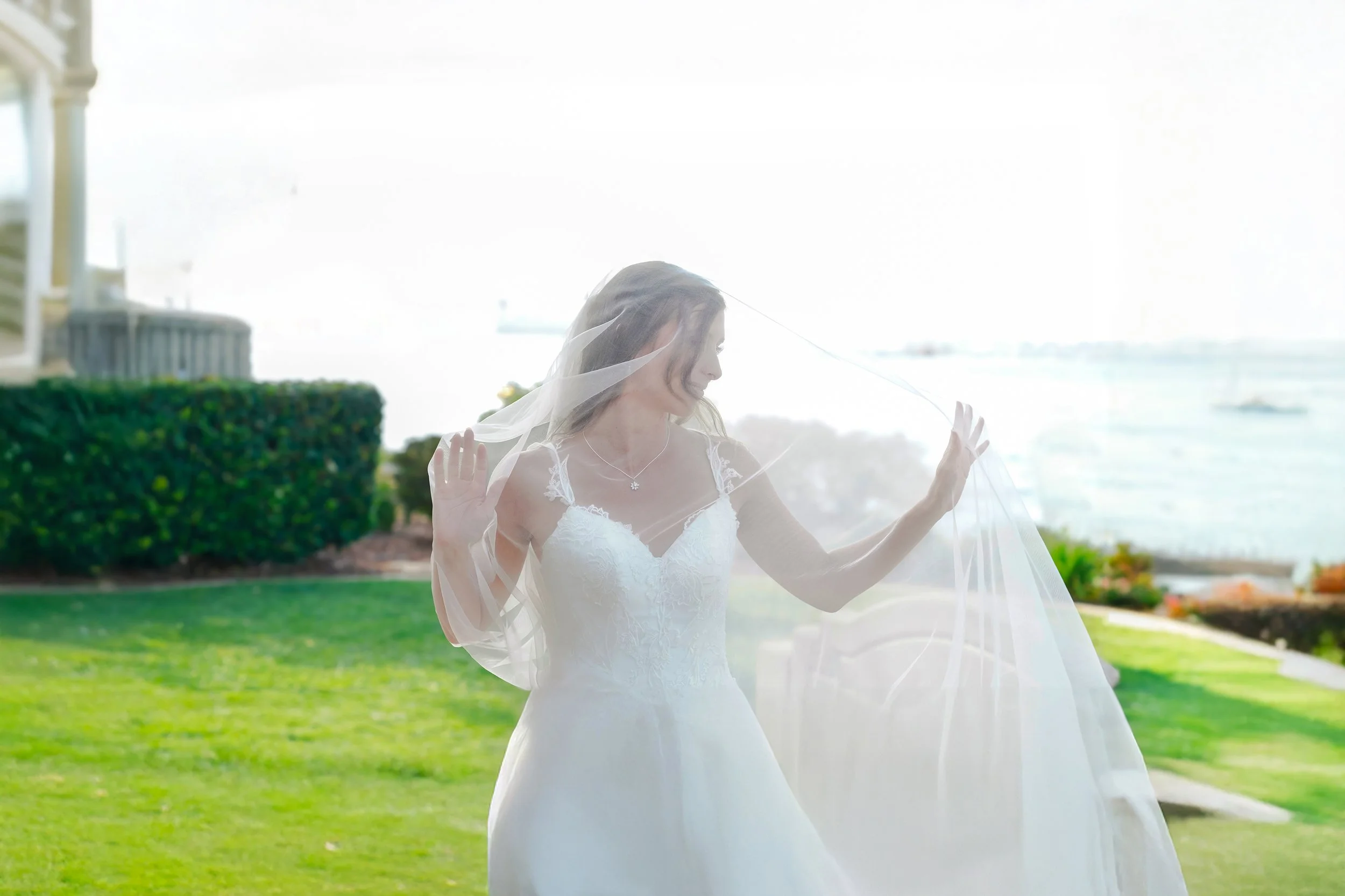 A bride wearing a white wedding dress and veil, standing outside near water, with her hands raised and looking down.