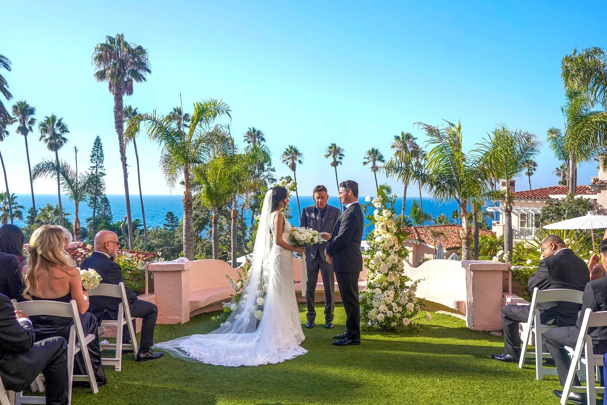 Outdoor wedding ceremony on the garden lawn at La Valencia Hotel in La Jolla with ocean views and floral arch pillars.