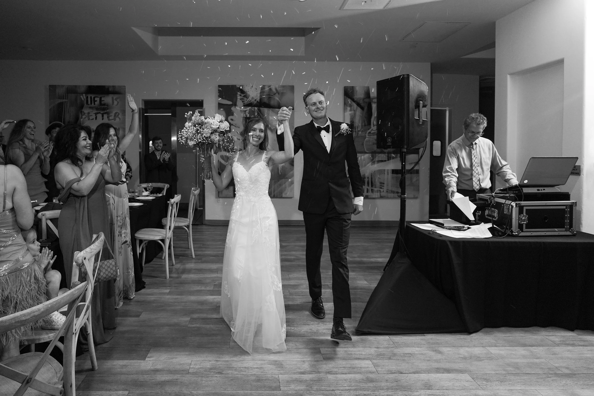 A newlywed couple, the bride holding a bouquet of flowers, walking hand in hand through a room filled with smiling wedding guests, some clapping. A DJ is visible on the right side with equipment. The room is decorated with artwork and a sign that rea