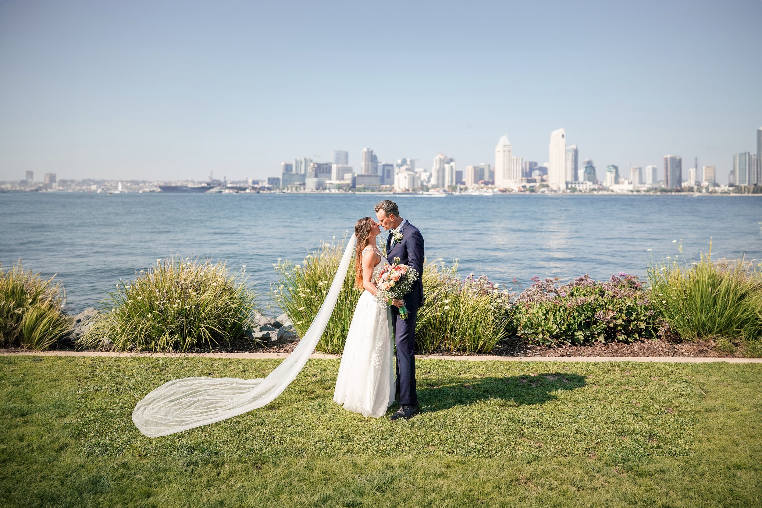 A bride and groom standing close to each other on a grassy area near the water with a city skyline in the background, holding a bouquet of flowers and touching foreheads.