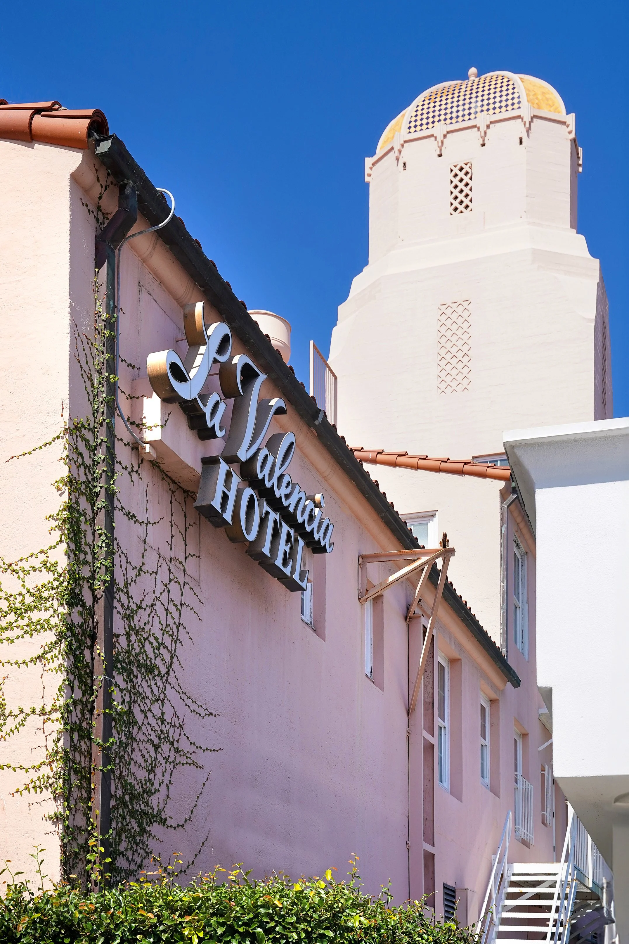 La Valencia Hotel exterior building sign on a pink stucco wall with green climbing ivy next to the historic Spanish revival tower under a blue sky in La Jolla.