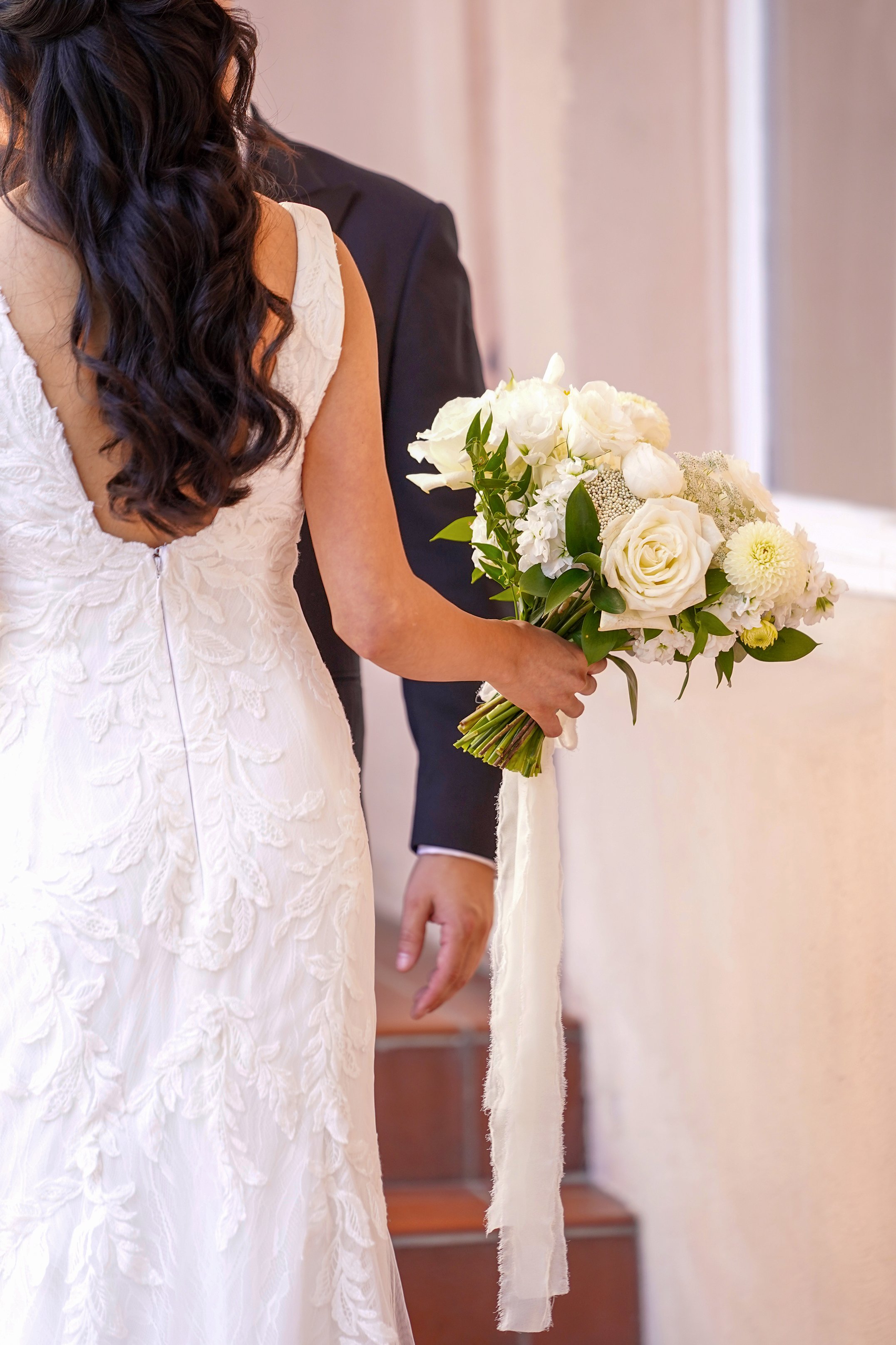 Rear view of a bride with long wavy half-up hair in a lace wedding dress holding a white rose bouquet with long silk ribbons.