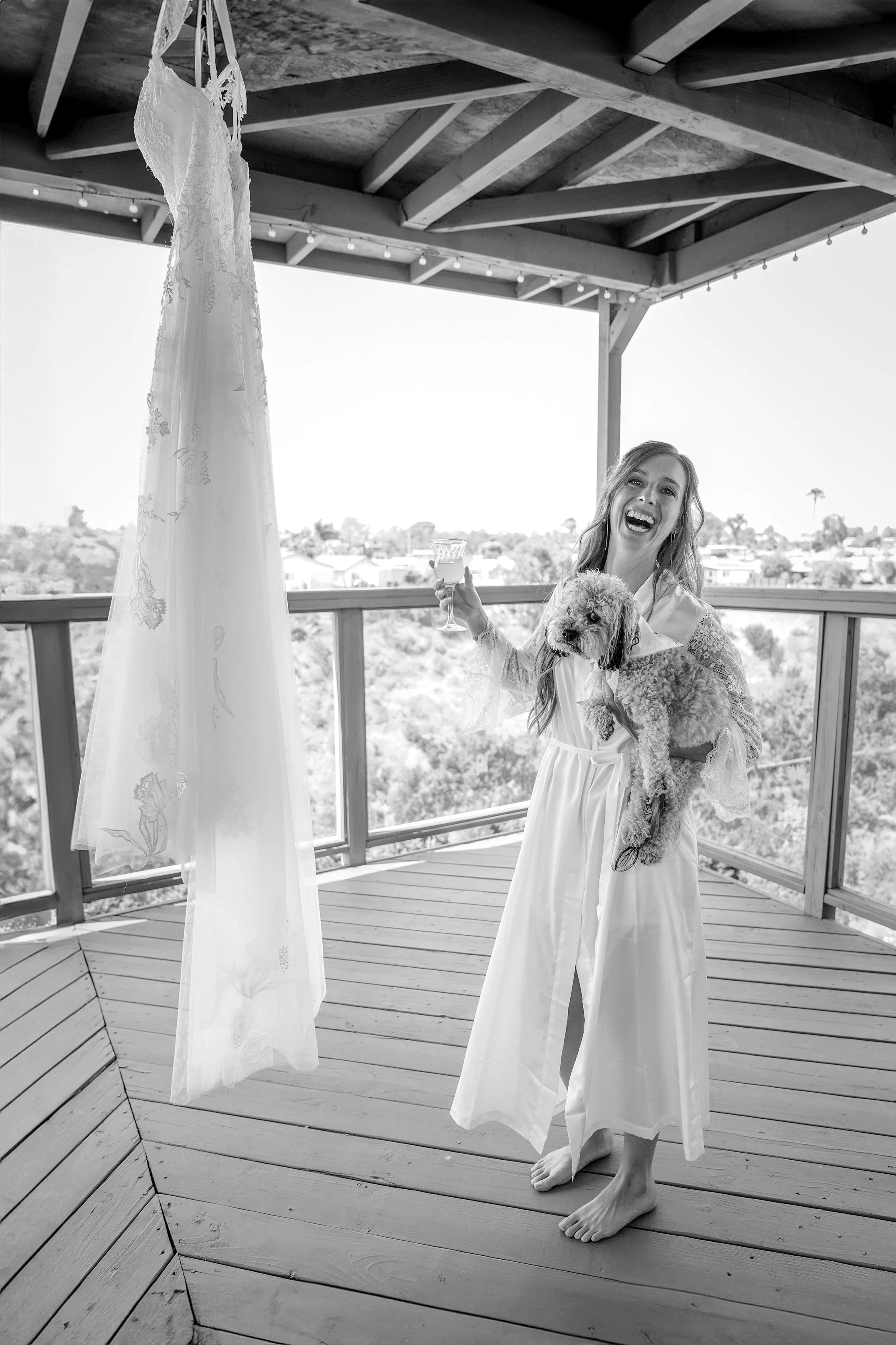 Woman holding a dog, standing barefoot on a wooden deck, wearing a white robe, with a wedding dress hanging from a ceiling post, outdoors under a roofed structure with string lights.