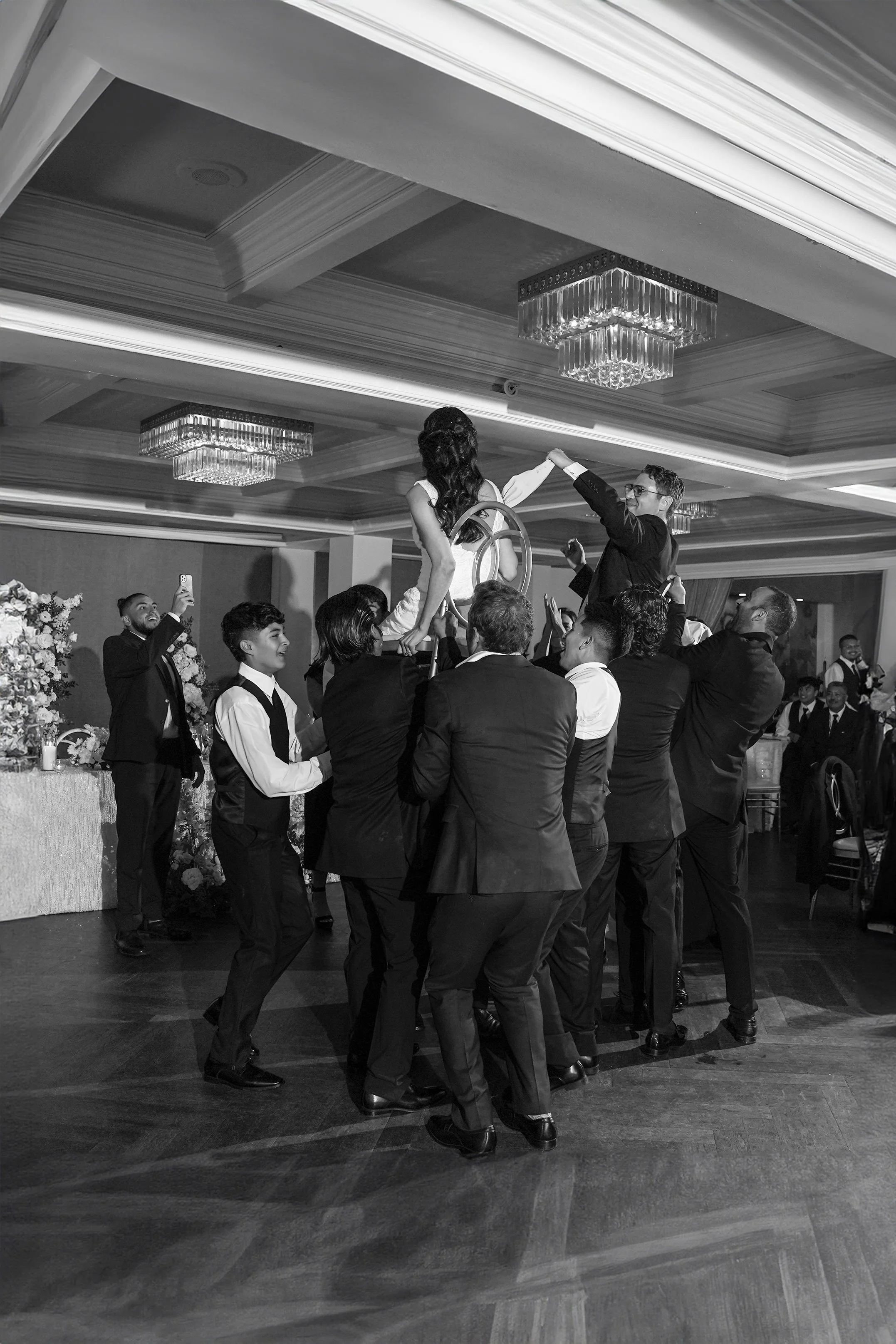 Black and white photo of a bride and groom being lifted on chairs during the Hora dance at their La Valencia Hotel wedding reception in La Jolla, San Diego.