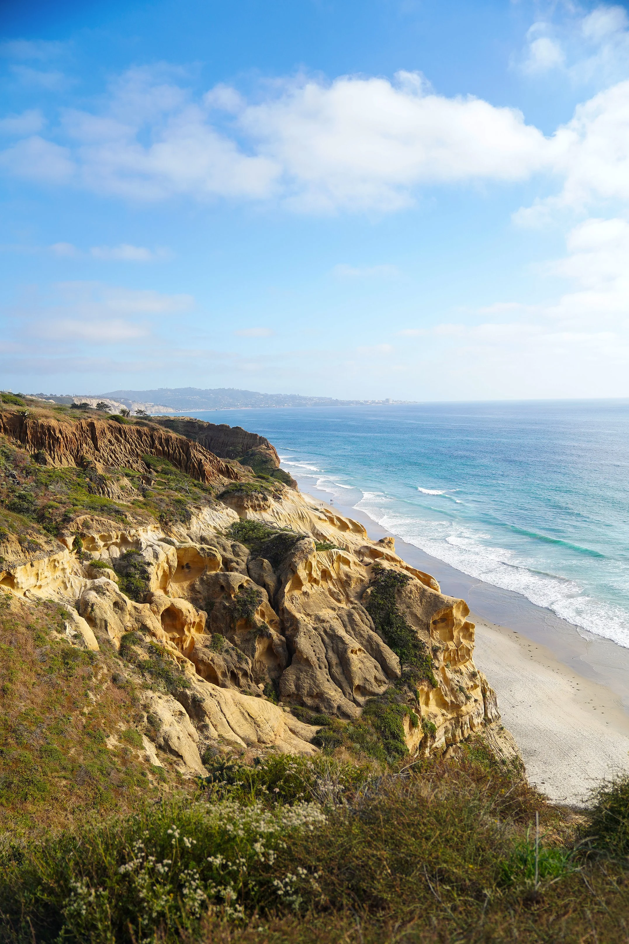 A high-angle, vertical shot captures a rugged coastal landscape where golden sandstone cliffs meet a sandy beach and a turquoise ocean under a blue sky with scattered clouds.