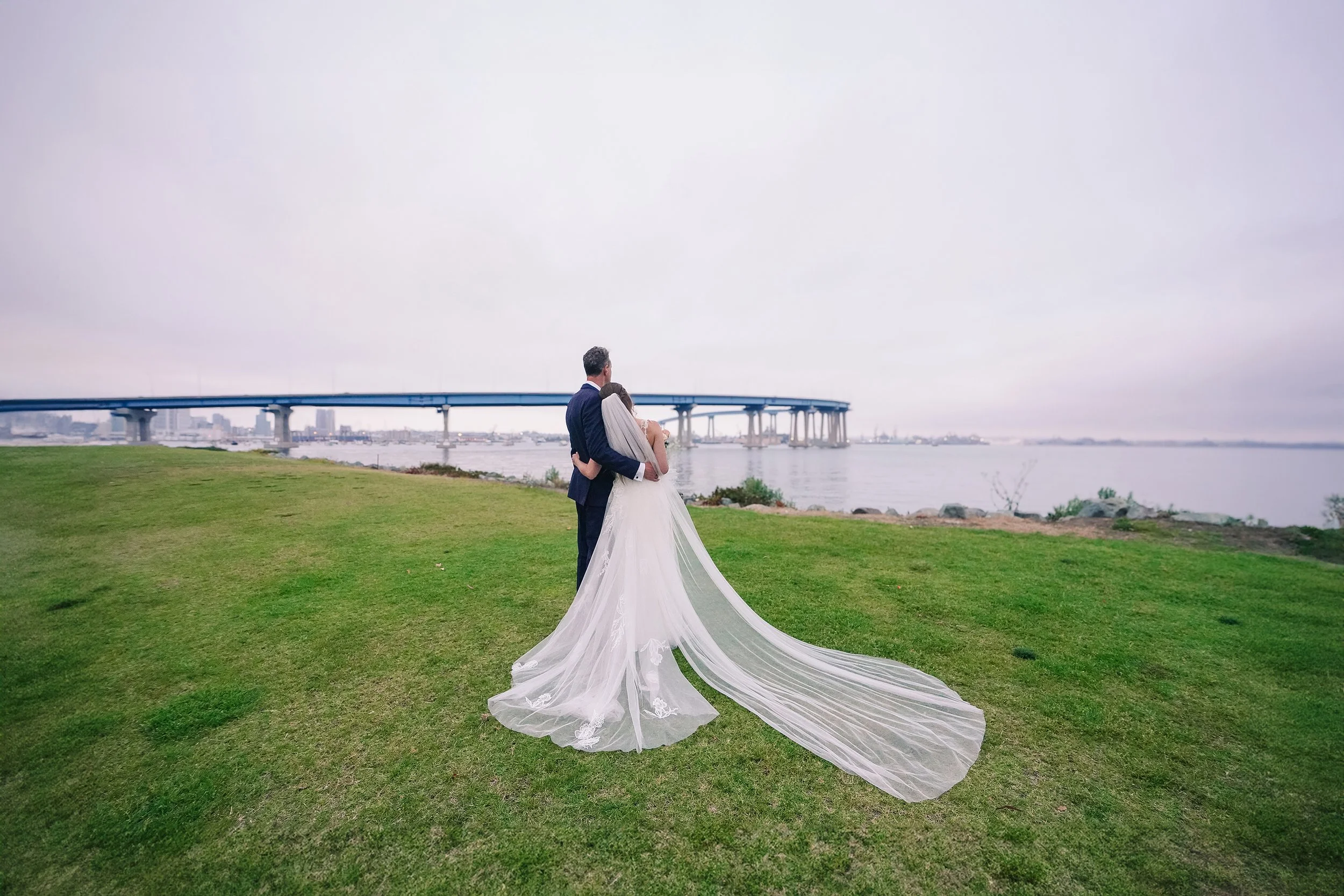 A wide shot from behind a bride in a white wedding gown and an ultra-long veil trailing on the grass, standing with her head on a groom's shoulder in a navy tuxedo. They are looking out over a calm bay toward the San Diego-Coronado Bay Bridge under a