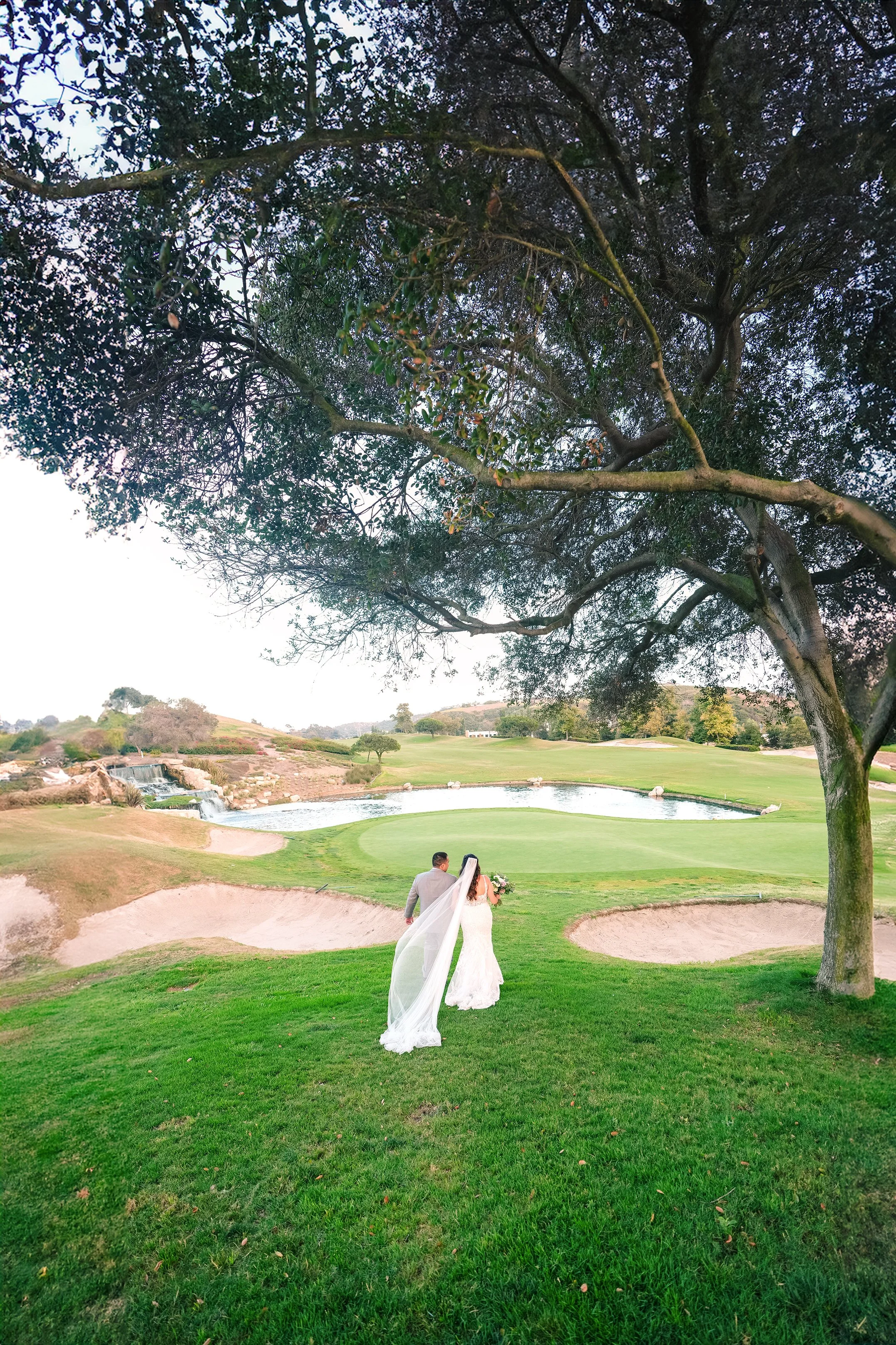 A bride and groom walk along the green golf course fairways under a large tree canopy during a wedding at the Crossing at Carlsbad.
