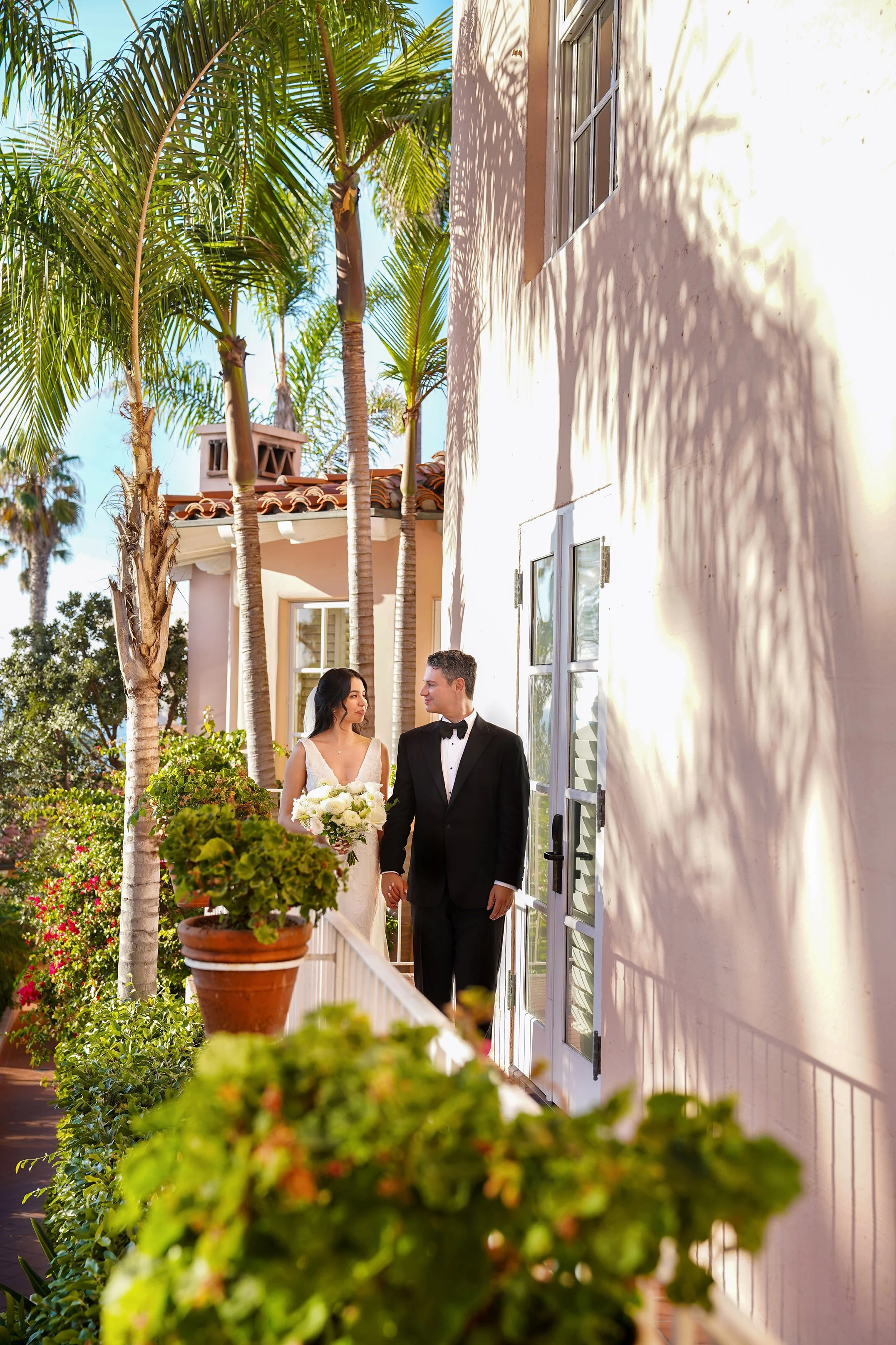 Bride and groom holding hands while walking on an outdoor terrace lined with palm trees and pink stucco walls during their wedding portraits at La Valencia Hotel in La Jolla, San Diego.